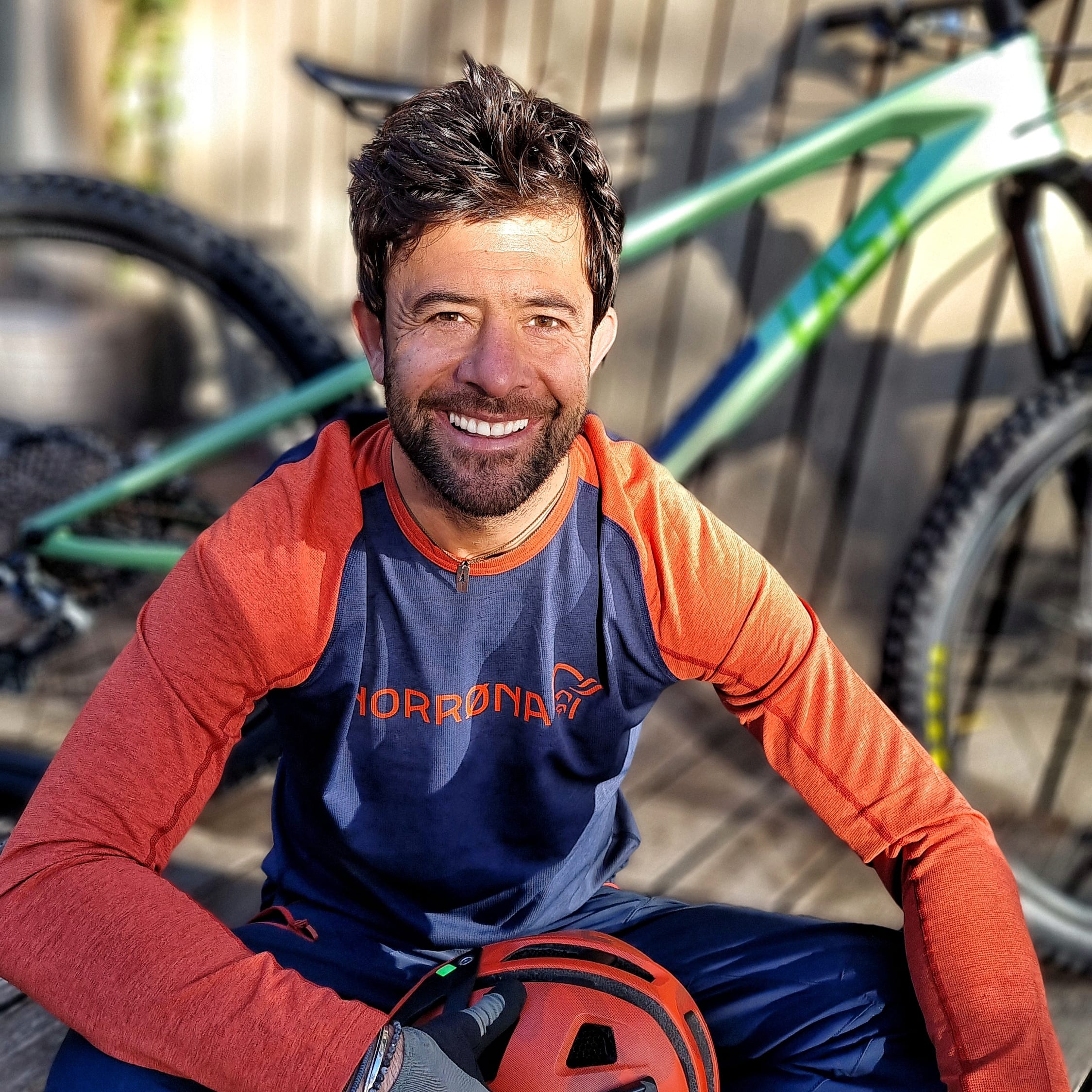 A man wearing an orange and blue long-sleeved shirt with a mountain logo is sitting on a wooden deck with his hands resting on his legs. He is holding a red bicycle helmet with a camera mount. A mountain bike is blurred in the background.