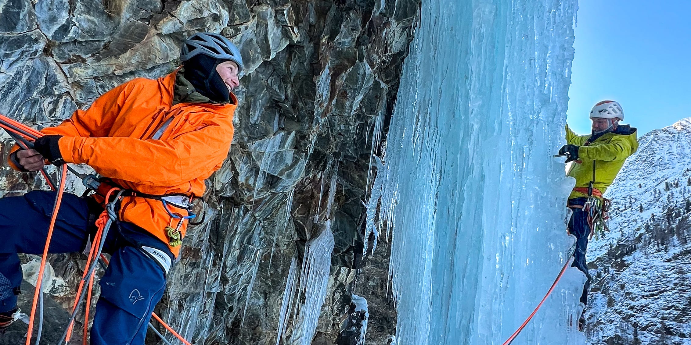 A climber wearing a Norrøna jacket and helmet is rappelling down an ice wall. A second climber is above. There are ropes attached to the climbers.