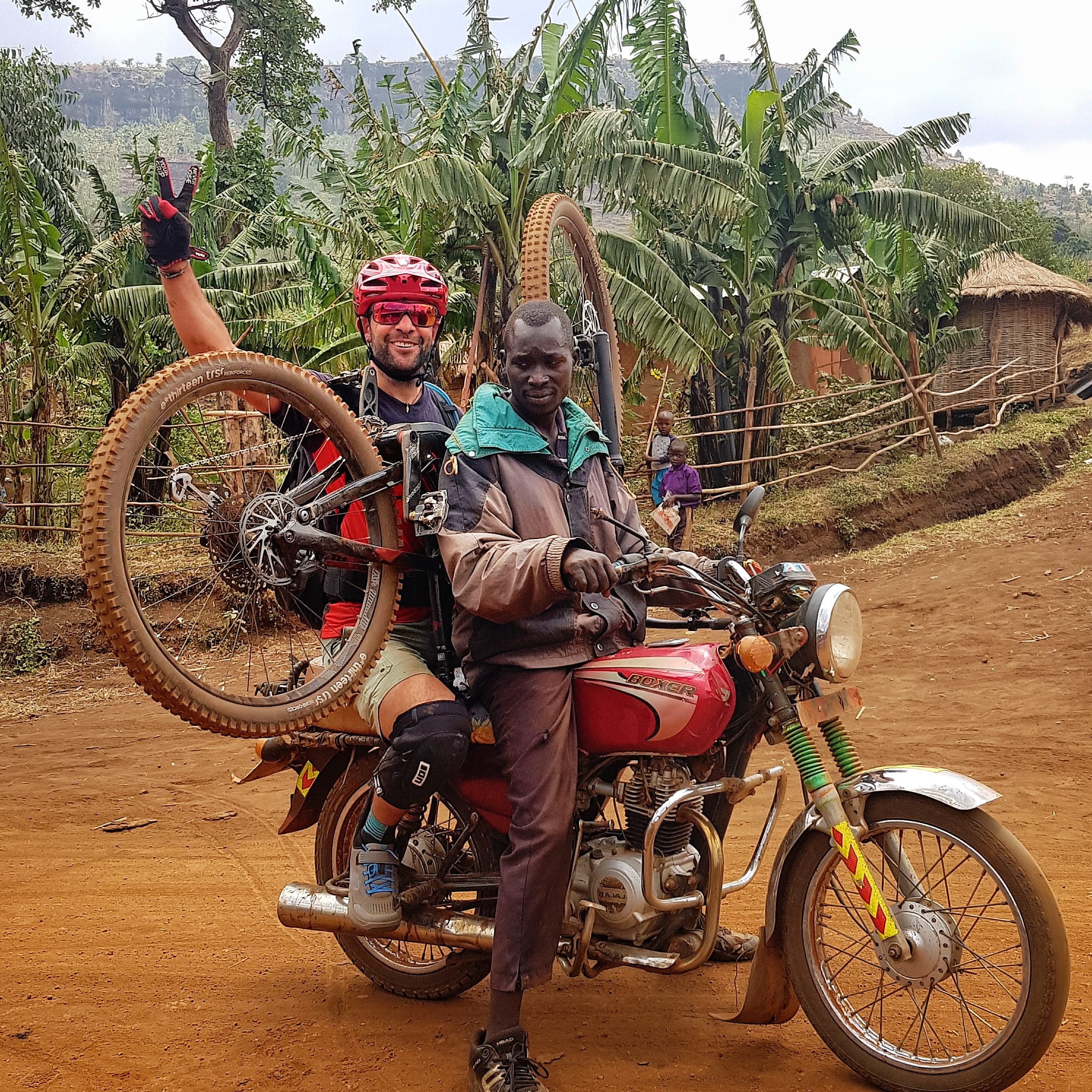 Two men, one wearing a red helmet and sunglasses, the other wearing a green jacket, are standing beside a red motorcycle. The man in the red helmet is holding a bicycle with the front wheel off, the man in the jacket is sitting on the motorcycle. The motorcycle is on a dirt road. 
