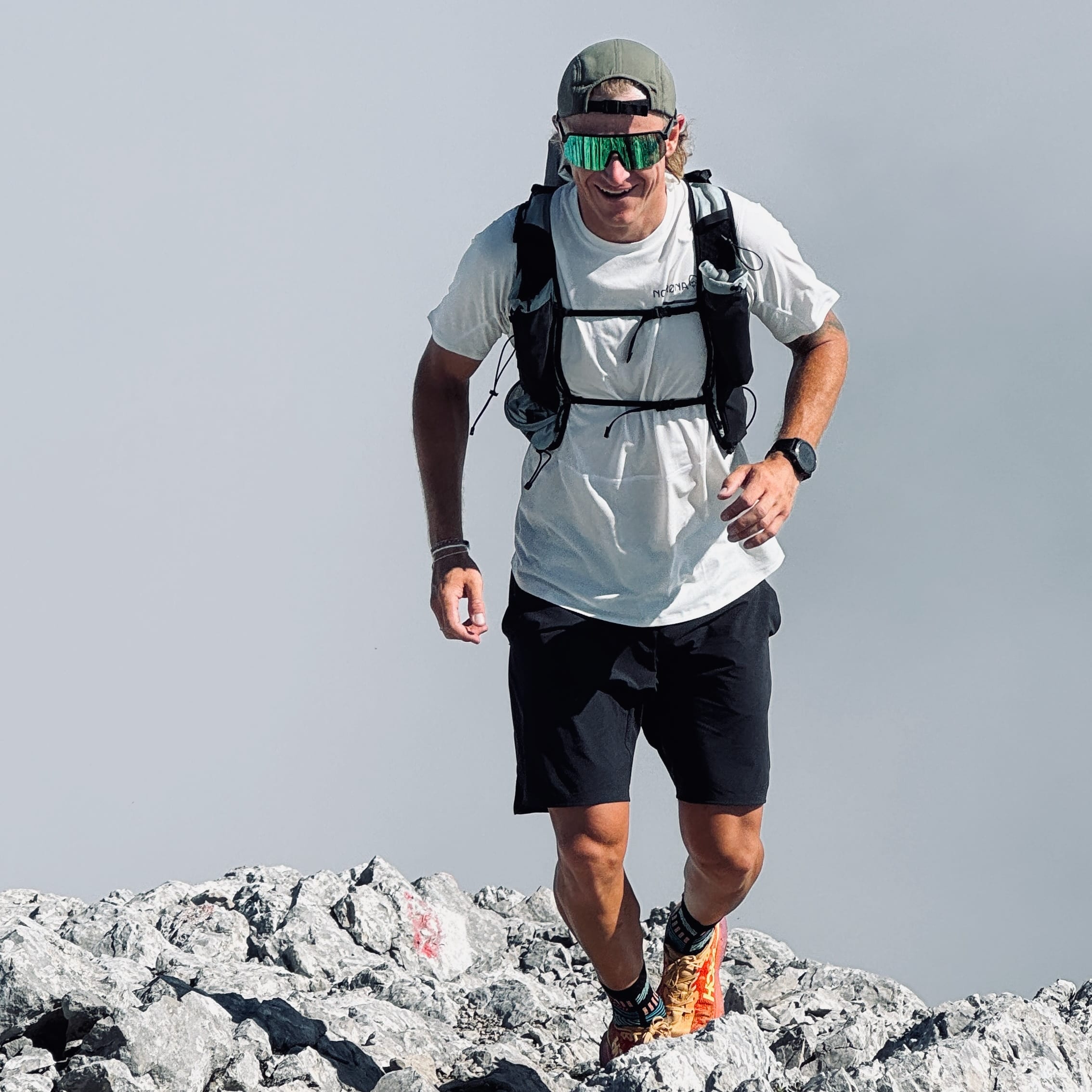 A man trail running on rocks, wearing a Norrøna hydration pack, t-shirt, shorts, and cap. He is smiling and wearing reflective sunglasses.