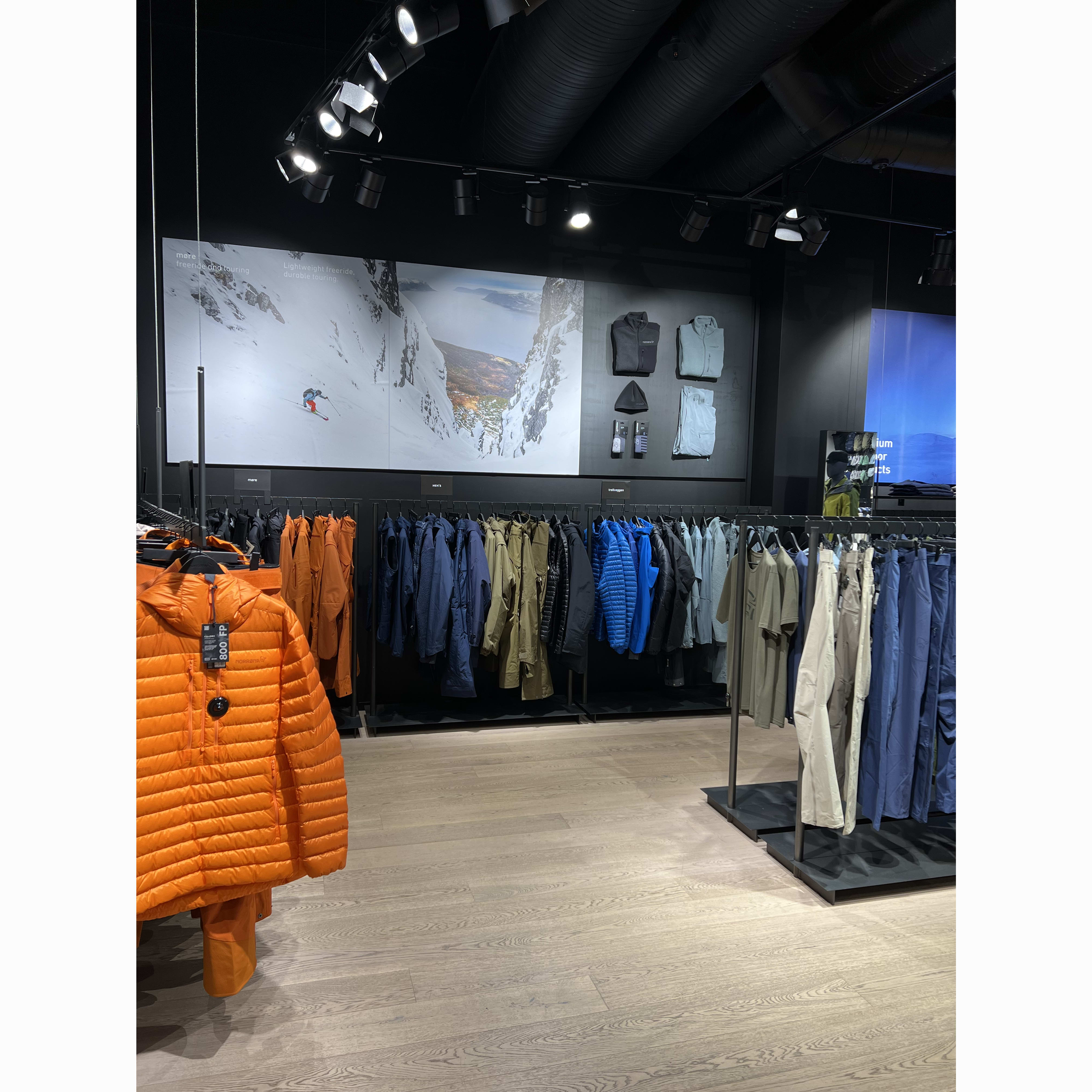 Retail store interior featuring rows of Norrøna apparel, including a prominent orange puffer jacket and a large mountain vista graphic.