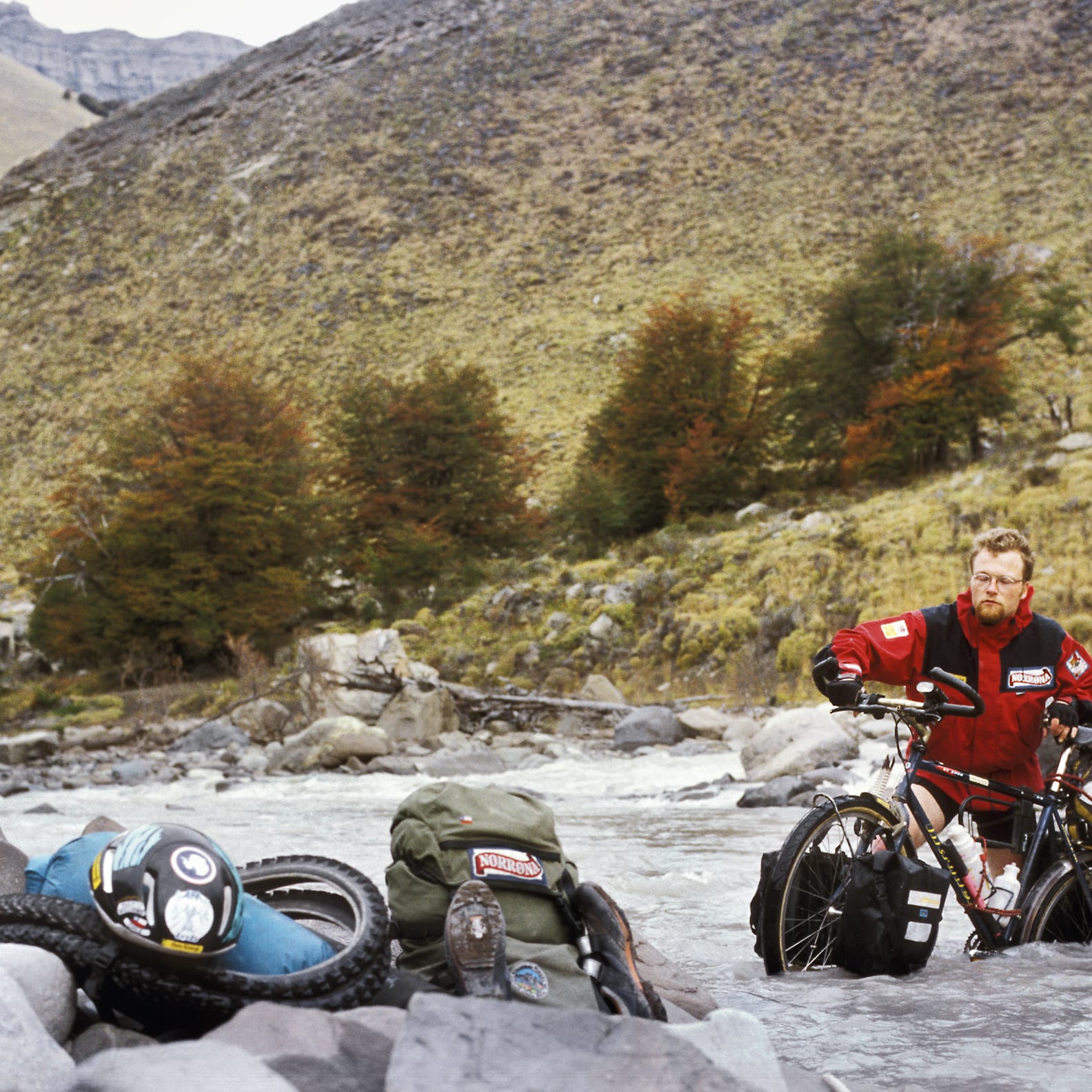 A man in a red jacket is riding a bicycle through a shallow river. He is wearing a helmet and has a backpack on his back. There are rocks and trees in the background.