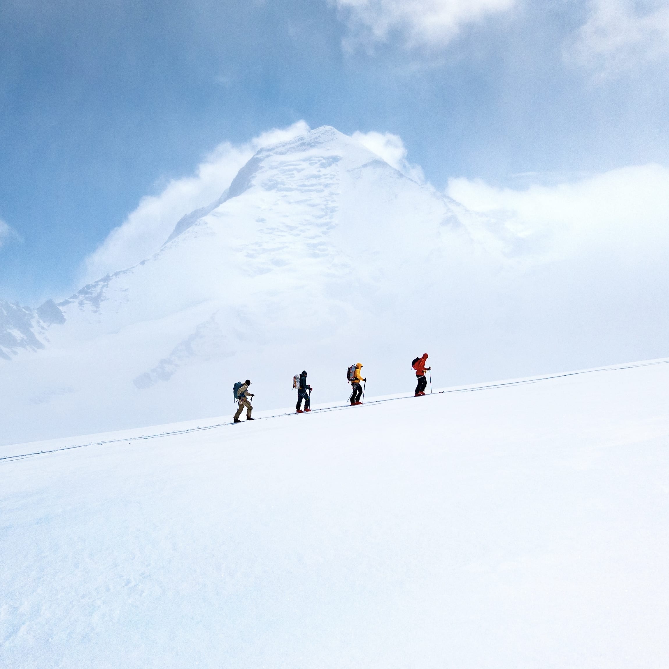 Four skiers ascend a snowy mountain with skis and poles, under a partly cloudy sky. The skier in orange is wearing Norrøna apparel.