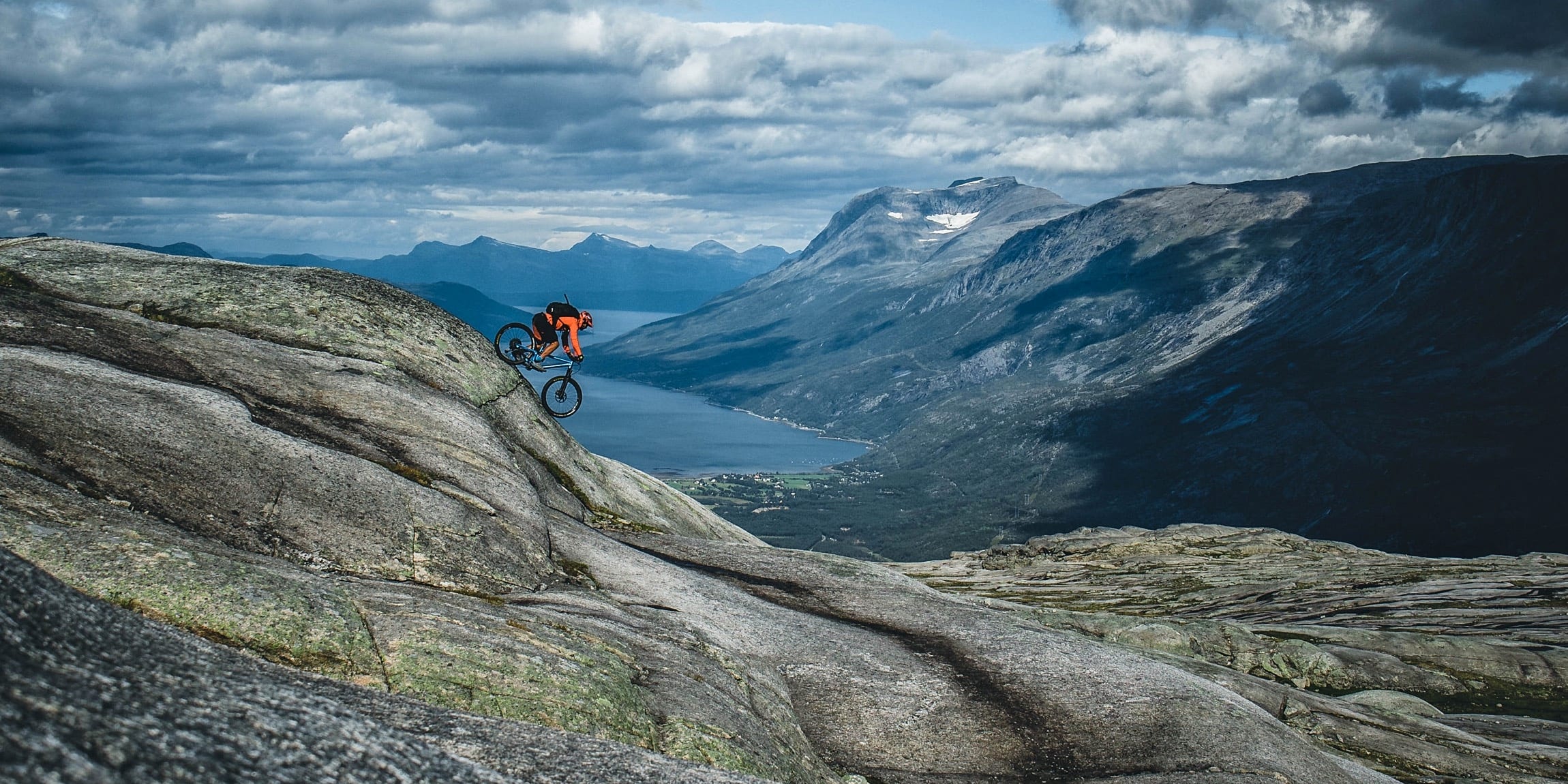 A mountain biker rides down a rocky trail with a valley and mountains in the background.  The biker is wearing an orange jacket and the trail is winding.