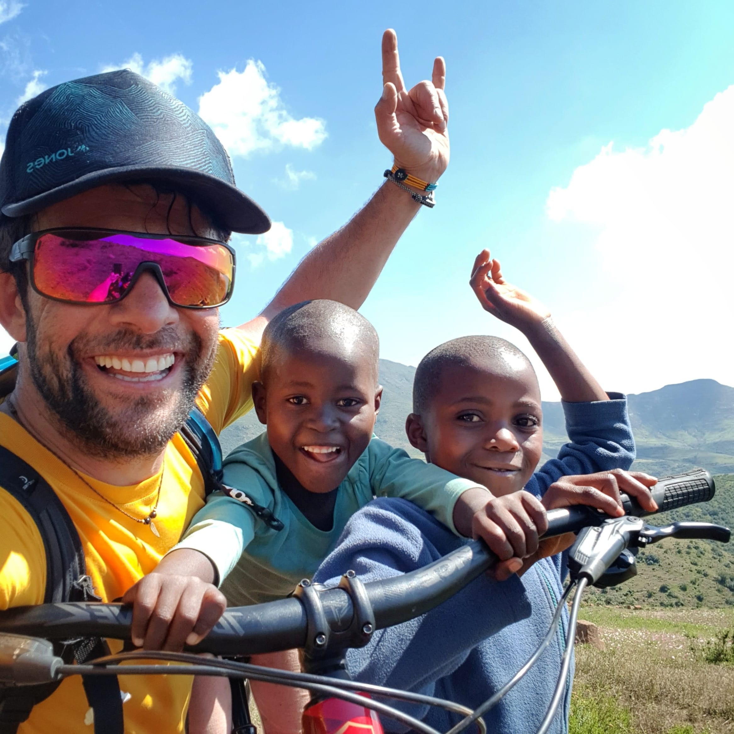 A man in a yellow shirt and sunglasses is smiling and holding a bicycle. Two children are smiling at the camera while sitting on the bike. The children are wearing blue and teal shirts.  They are outdoors in a grassy area, with mountains in the background.