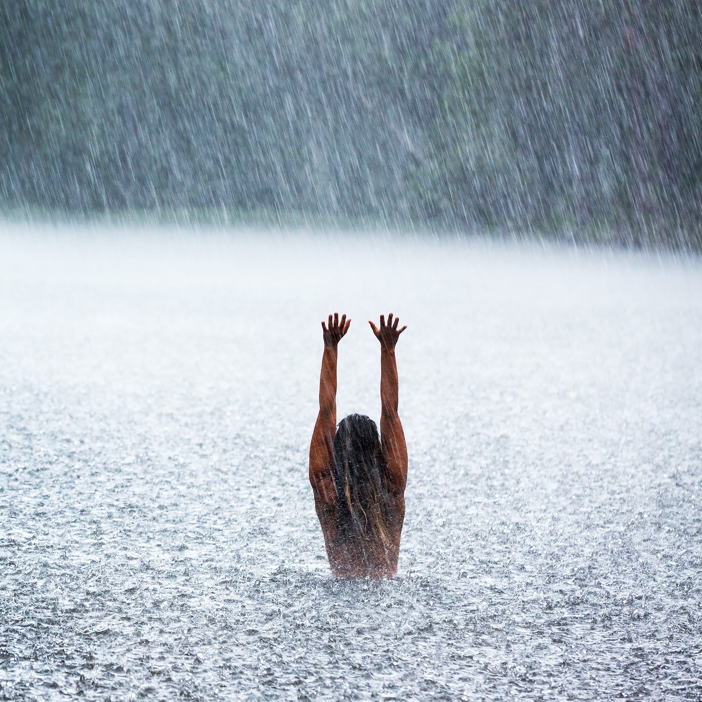 A person with arms raised, standing in a lake during a heavy rain shower. The person's head and shoulders are submerged in the water.