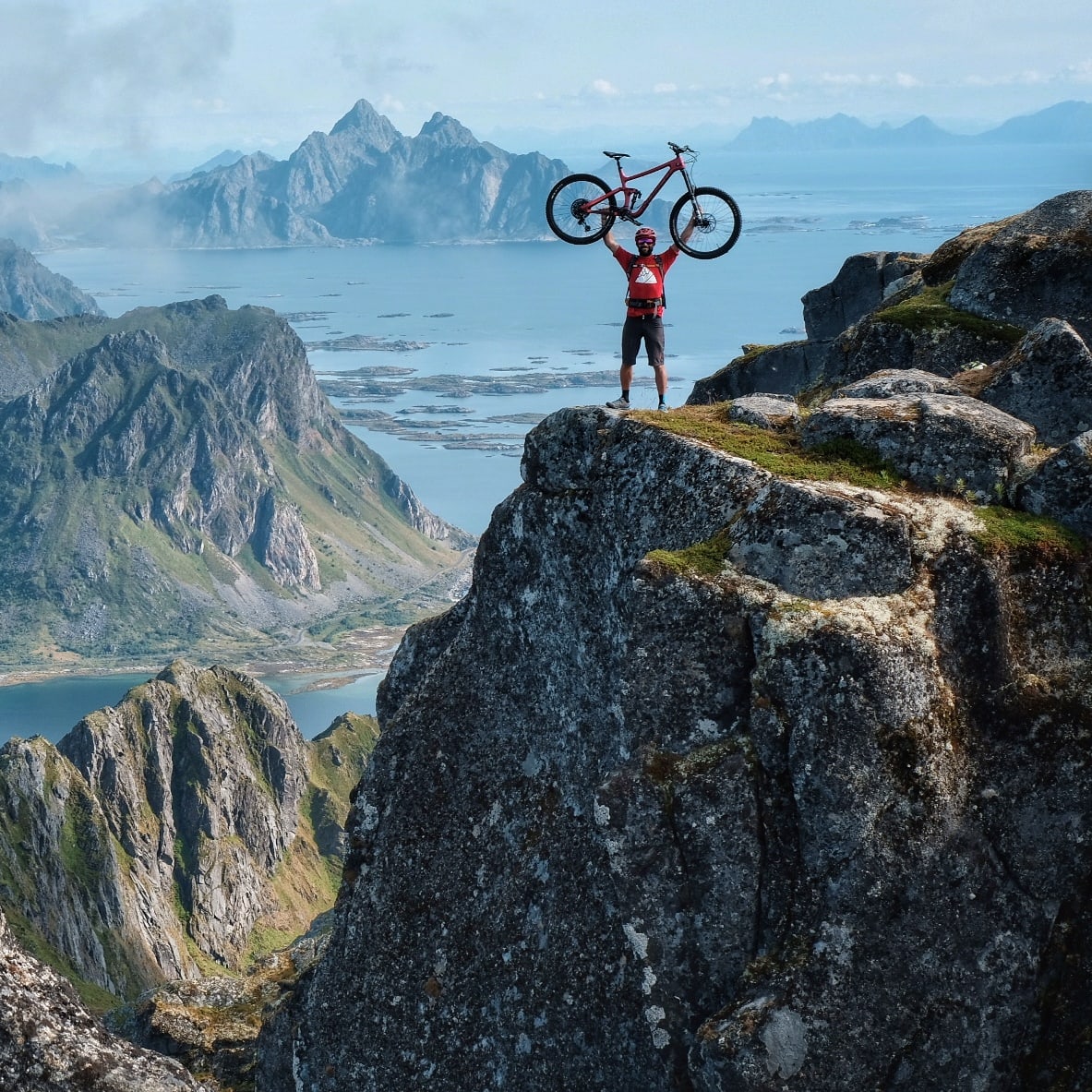 A person in Norrøna clothing stands on a mountaintop, holding a bicycle above their head, with a view of a fjord and mountains. 
