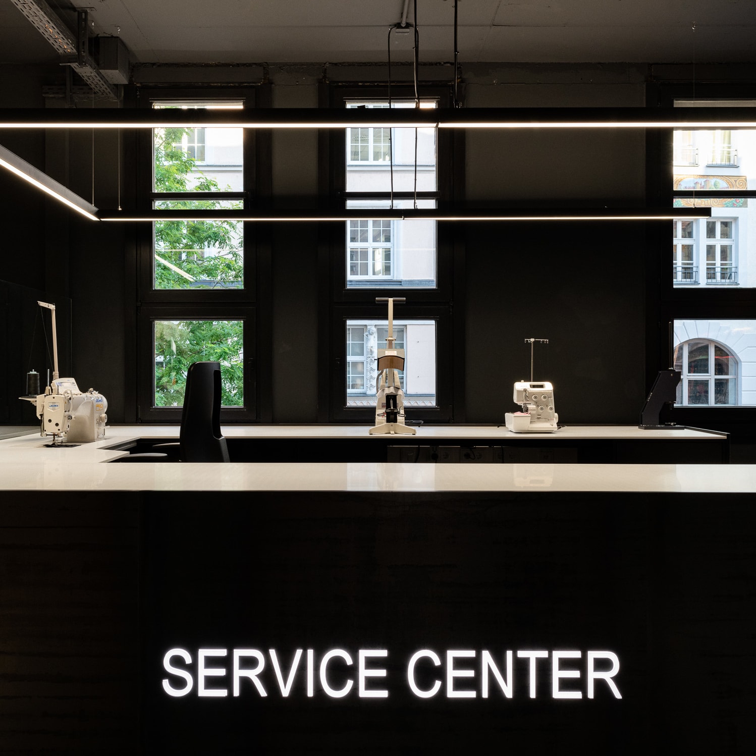 A dark room with large windows, a white counter with "SERVICE CENTER" in white lights, and two sewing machines. 

