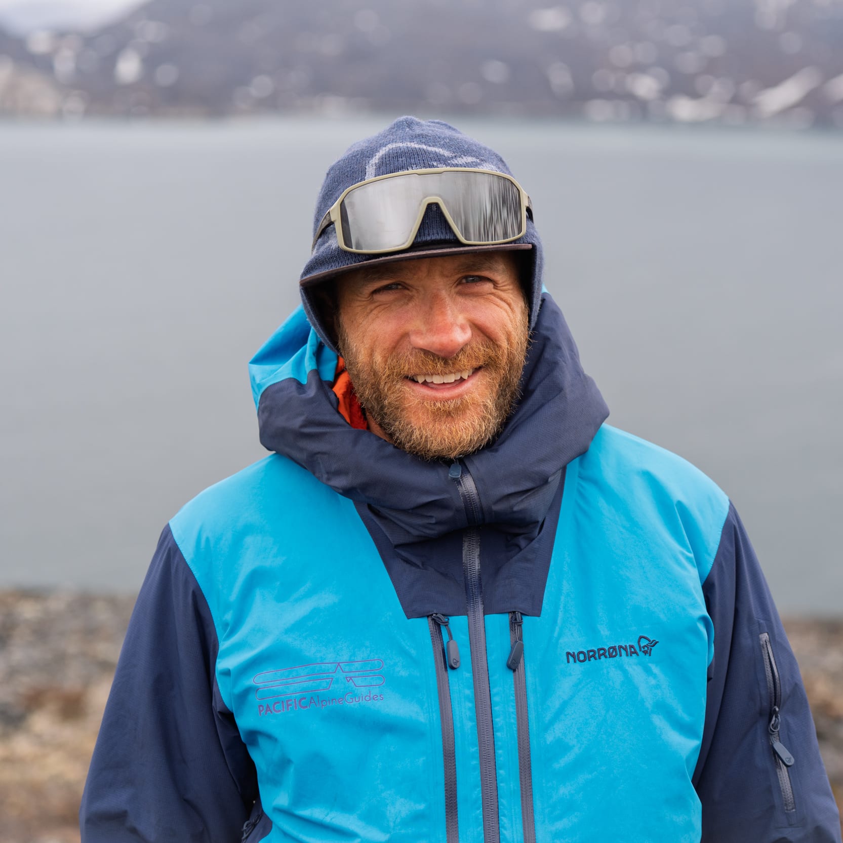 A smiling person with a beard, wearing a blue Norrøna jacket and sunglasses, stands in front of a body of water and distant snowy mountains.
