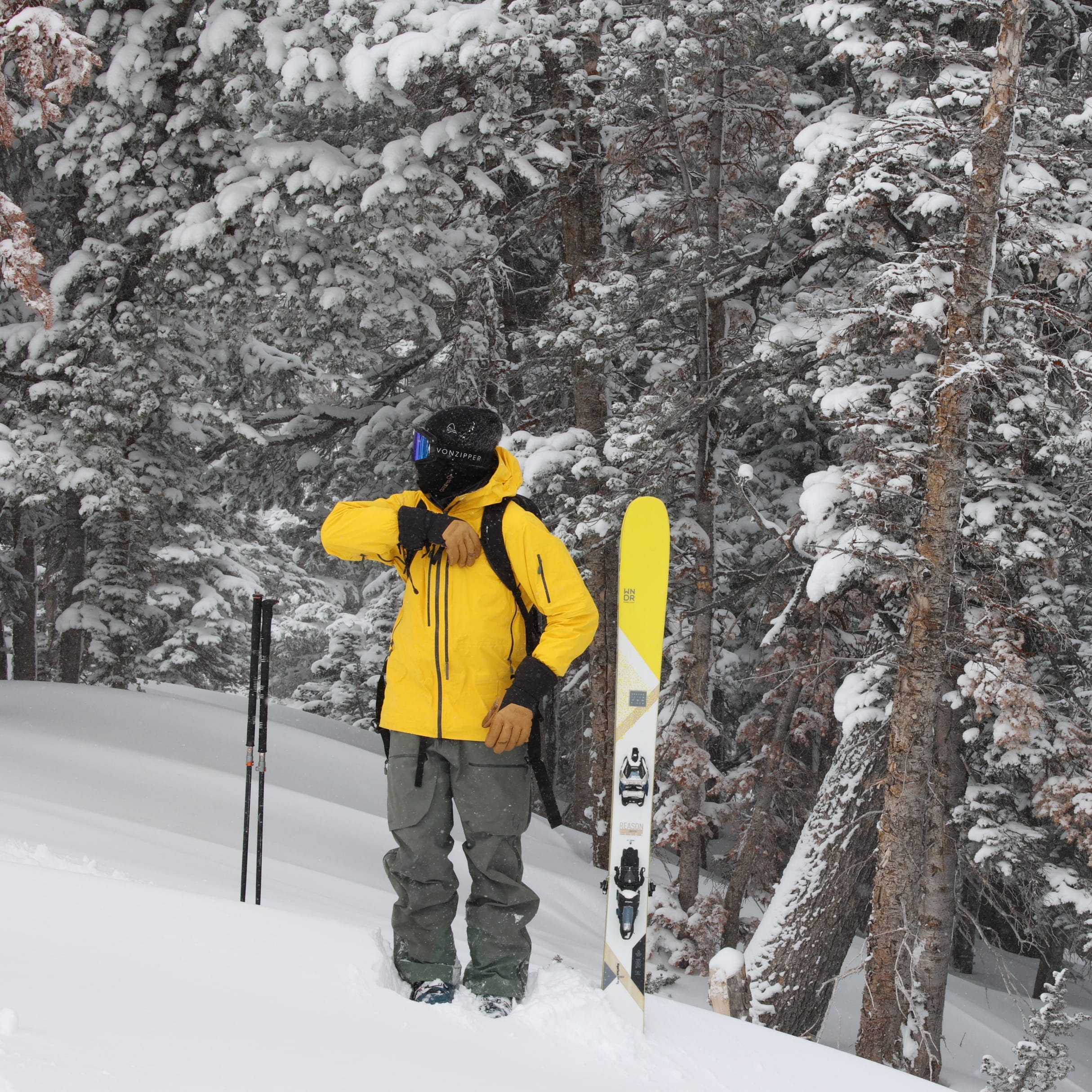 A person wearing a yellow Norrøna jacket and ski goggles standing in the snow next to a pair of skis in a forest.