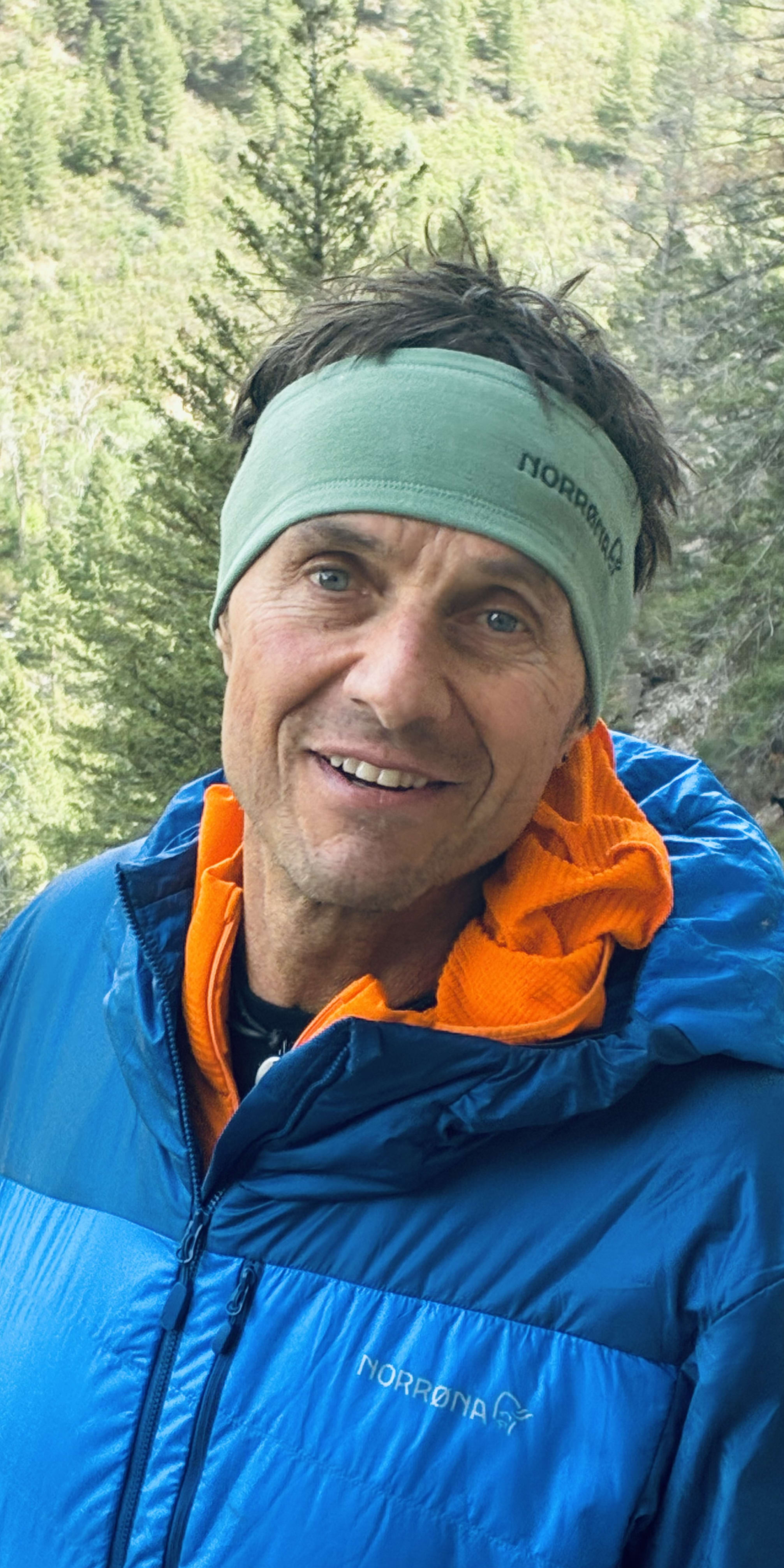 Smiling man outdoors, wearing a blue Norrøna jacket and green Norrøna headband. Orange shirt visible. Green trees in background.