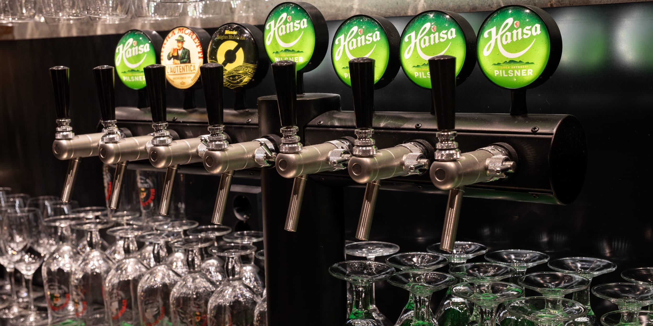 Close-up of a bar with rows of beer taps with "Hansa" and other logos, with rows of clean glasses underneath.