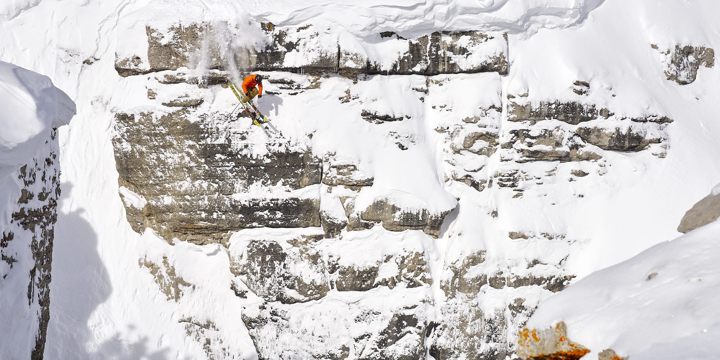 A skier in a Norrøna jacket jumps a snowy cliff, kicking up snow. The cliff is layered with rock and snow.