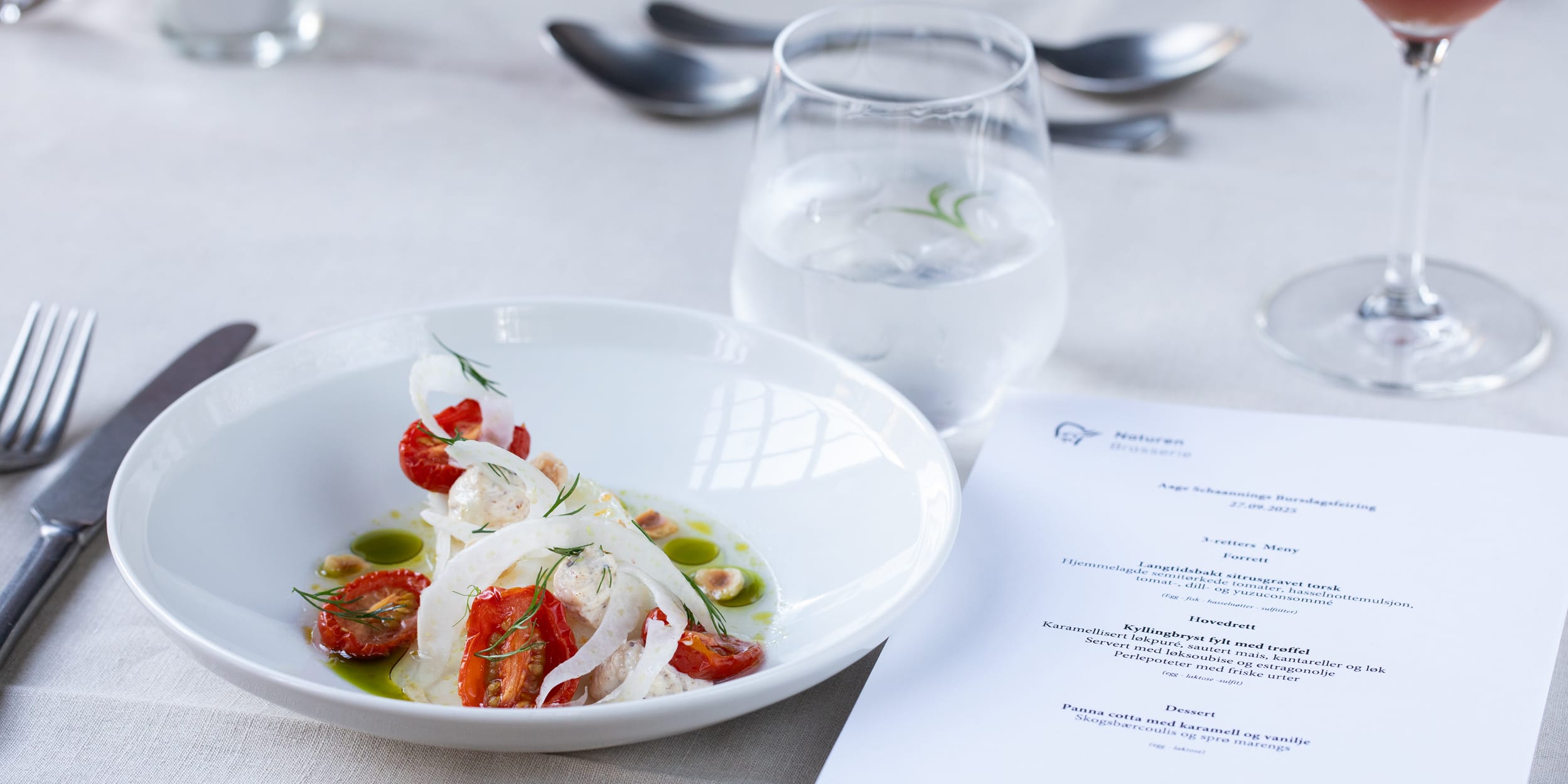A prepared meal of sliced tomatoes and mozzarella on a white plate with a menu, silverware, glass of water, wine glass, and candle on a table.