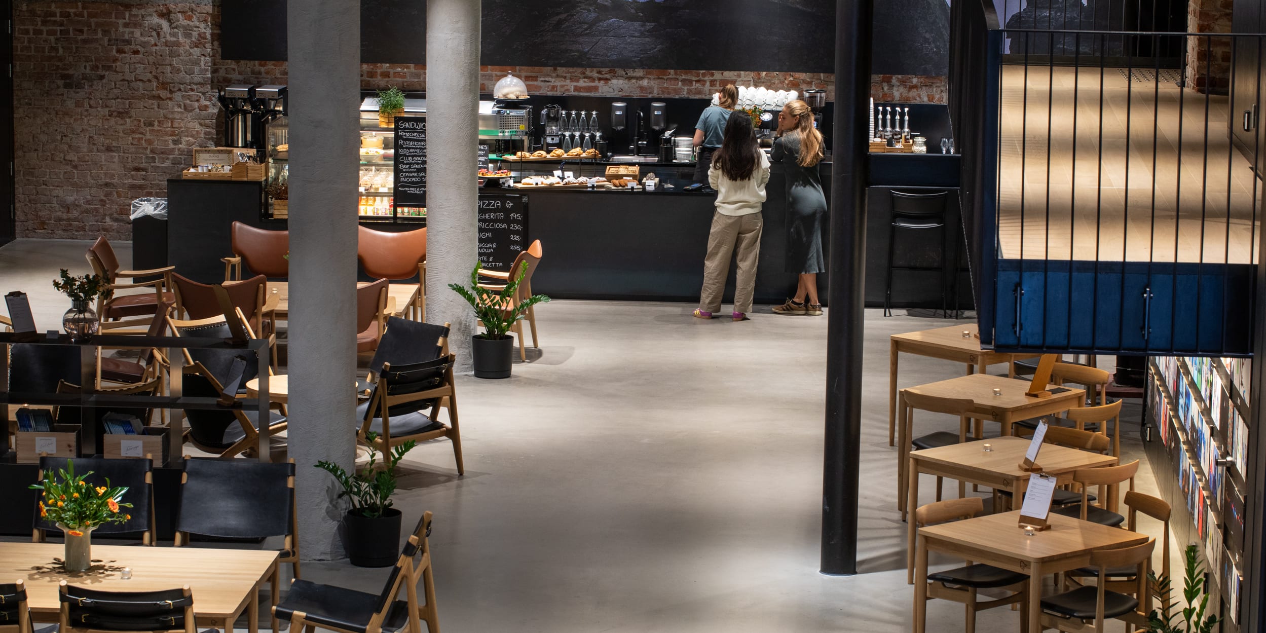 Interior of a cafe with brick walls, concrete columns, and wooden furniture. People are at the counter with baked goods. The person on the right is wearing Norrøna clothing.