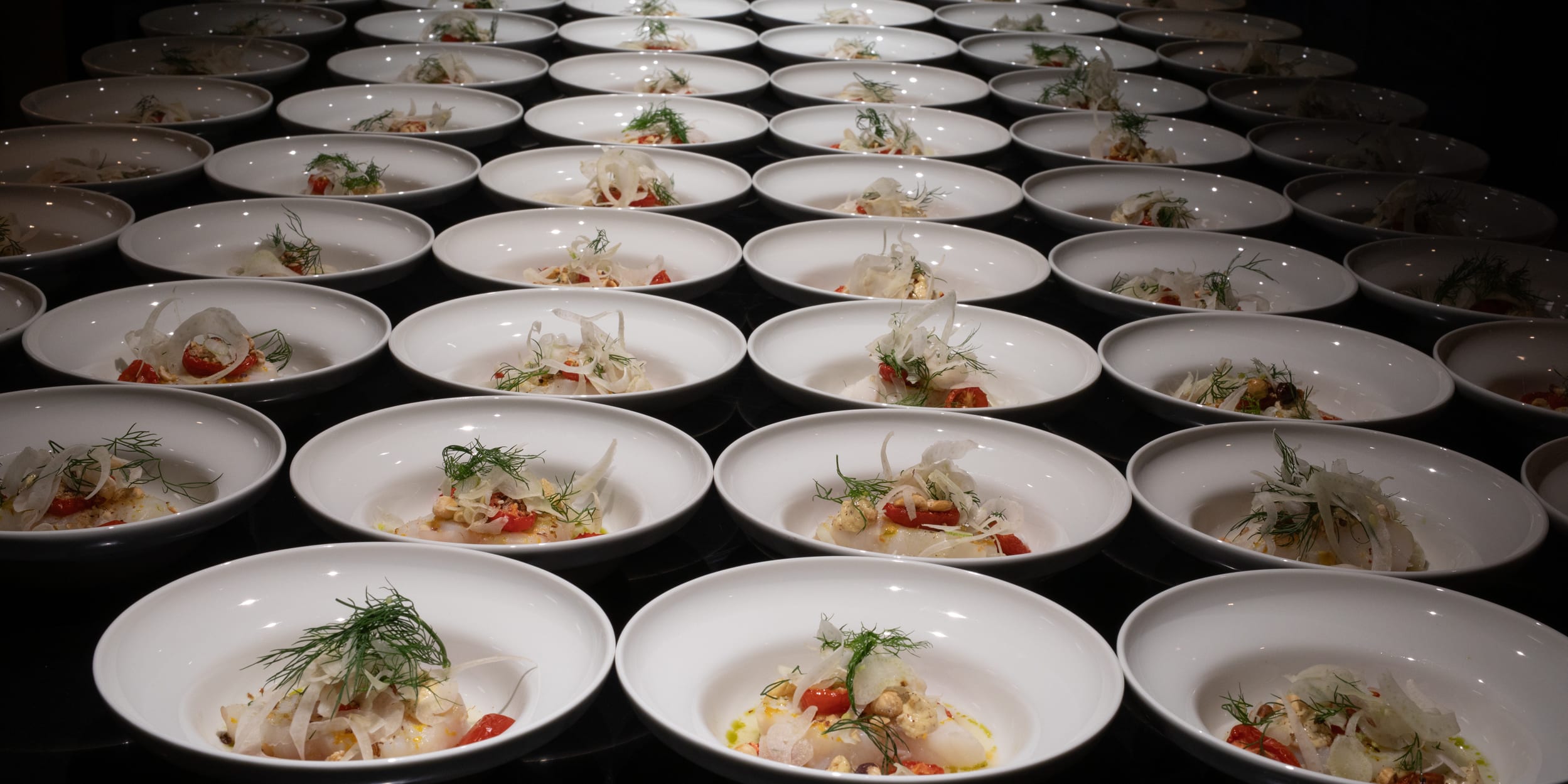 Rows of white plates hold a dish featuring white fish, sliced tomatoes, fennel, and dill, illuminated by overhead lighting.