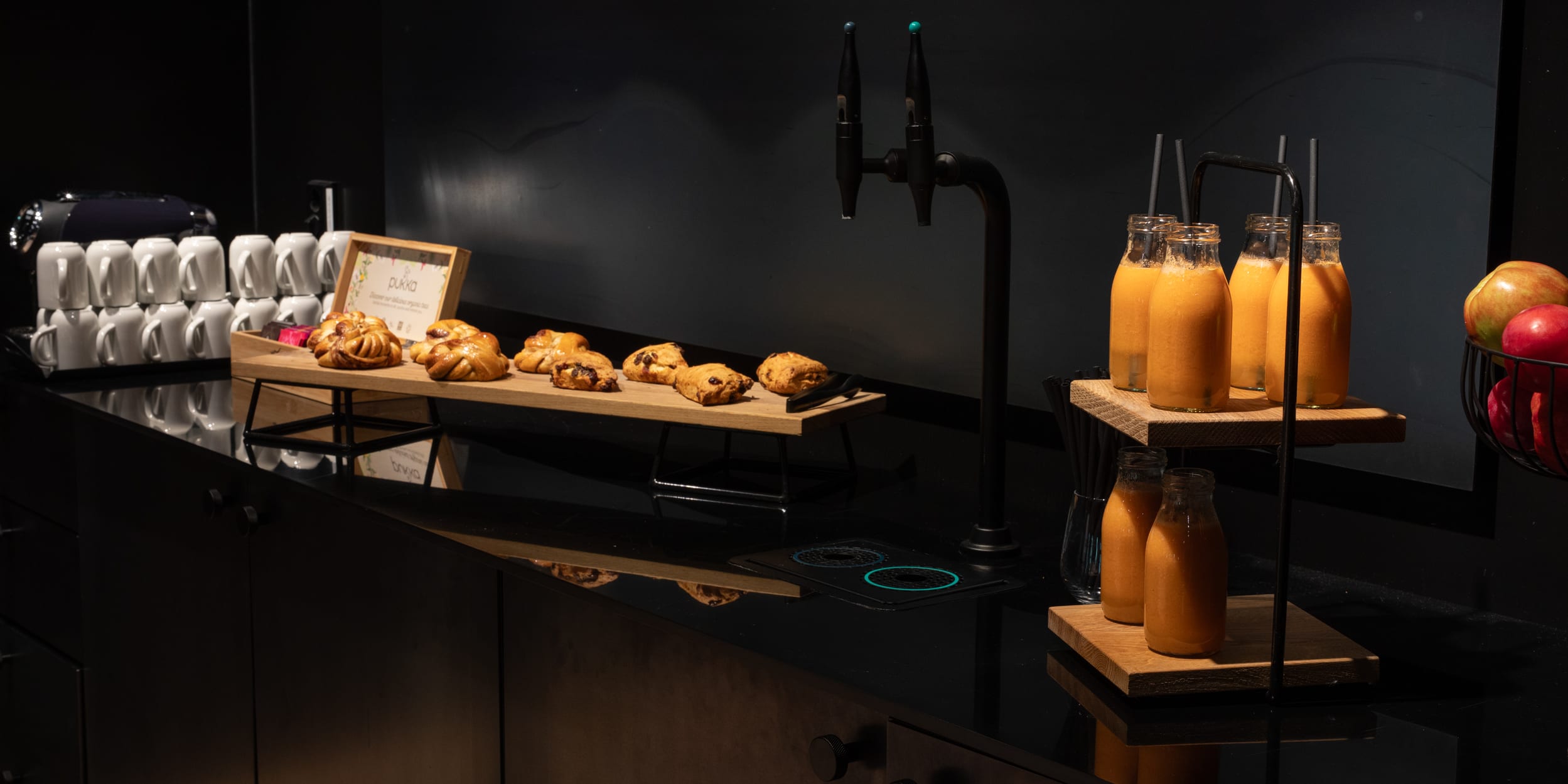 A breakfast buffet with pastries on a wooden platter, juice bottles with straws, stacked mugs, and a fruit basket against a dark background.