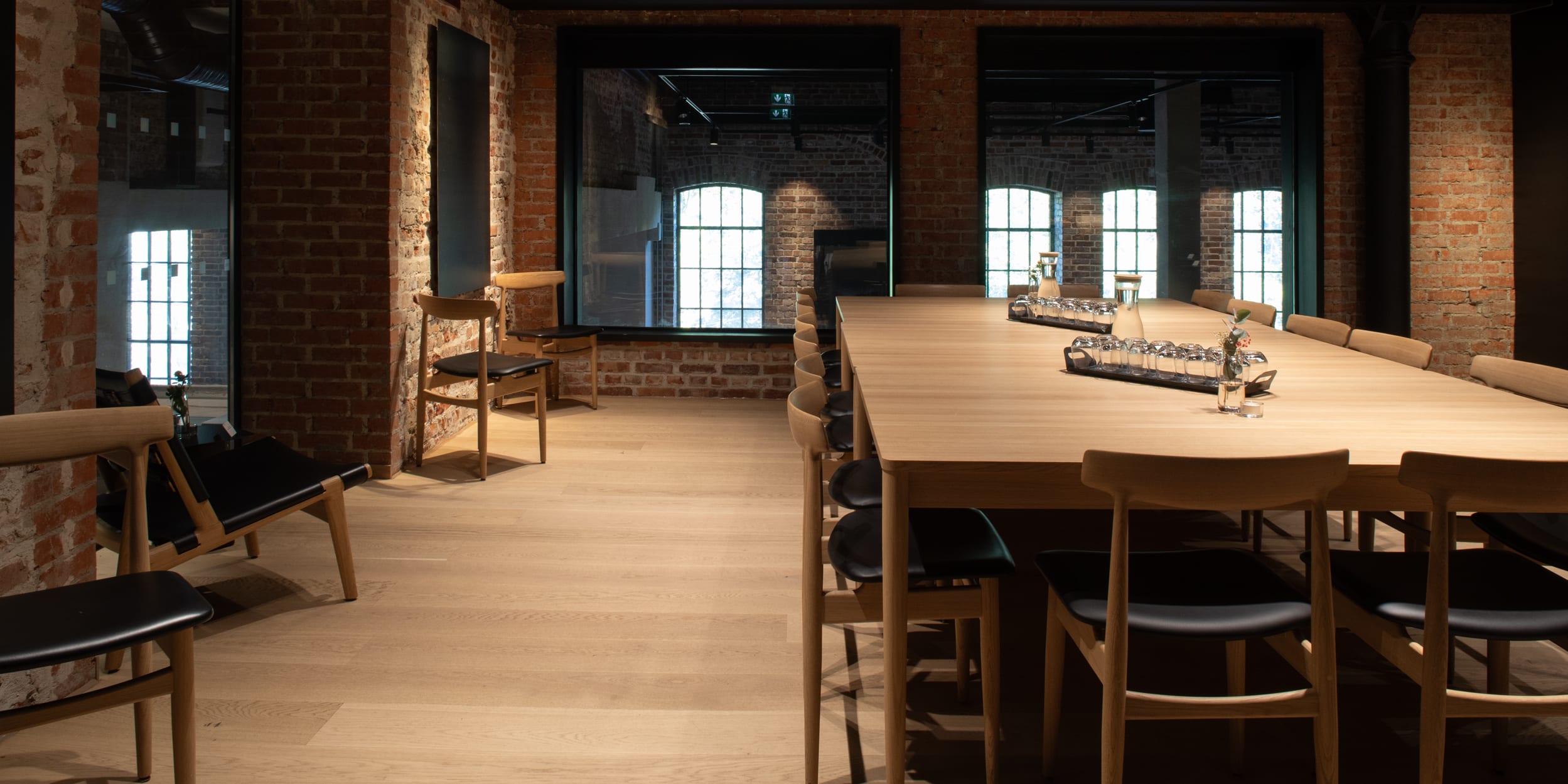 Conference room with brick walls, wood floors, a long table with chairs, and windows reflecting an industrial setting.