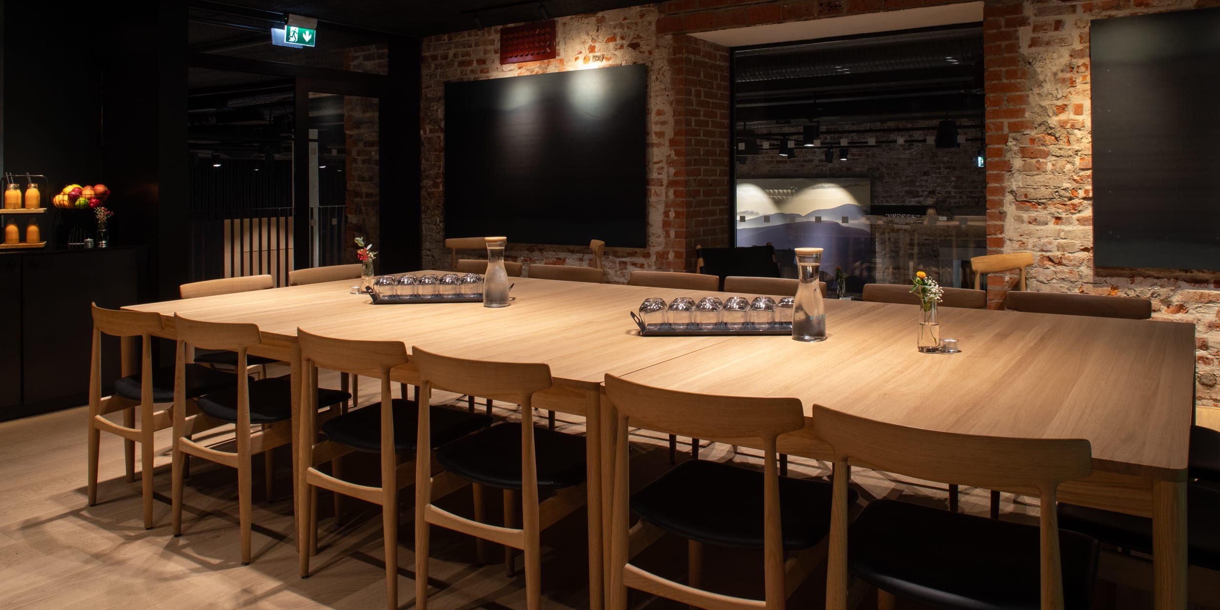 A long wooden table with chairs in a modern meeting room with exposed brick walls. There are carafes and glasses on the table.