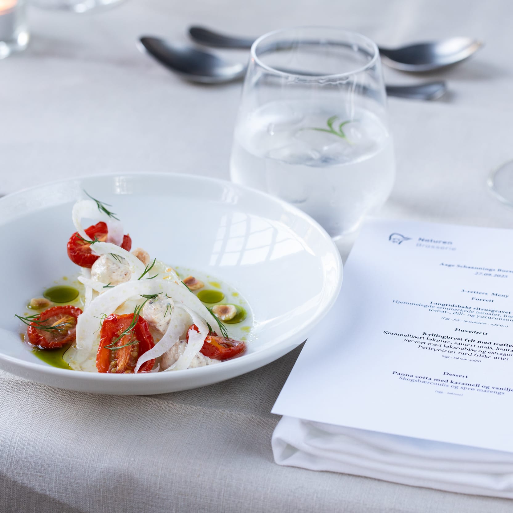A prepared meal of sliced tomatoes and mozzarella on a white plate with a menu, silverware, glass of water, wine glass, and candle on a table.