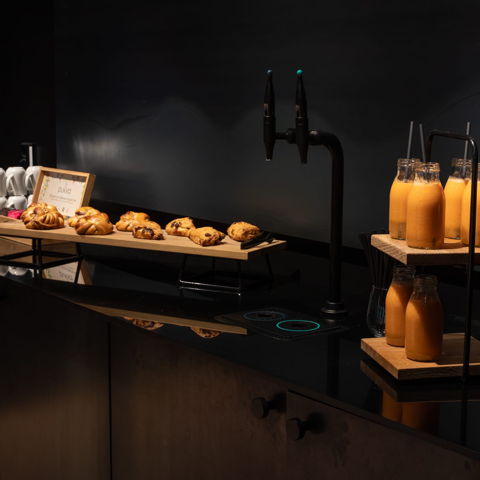 A breakfast buffet with pastries on a wooden platter, juice bottles with straws, stacked mugs, and a fruit basket against a dark background.