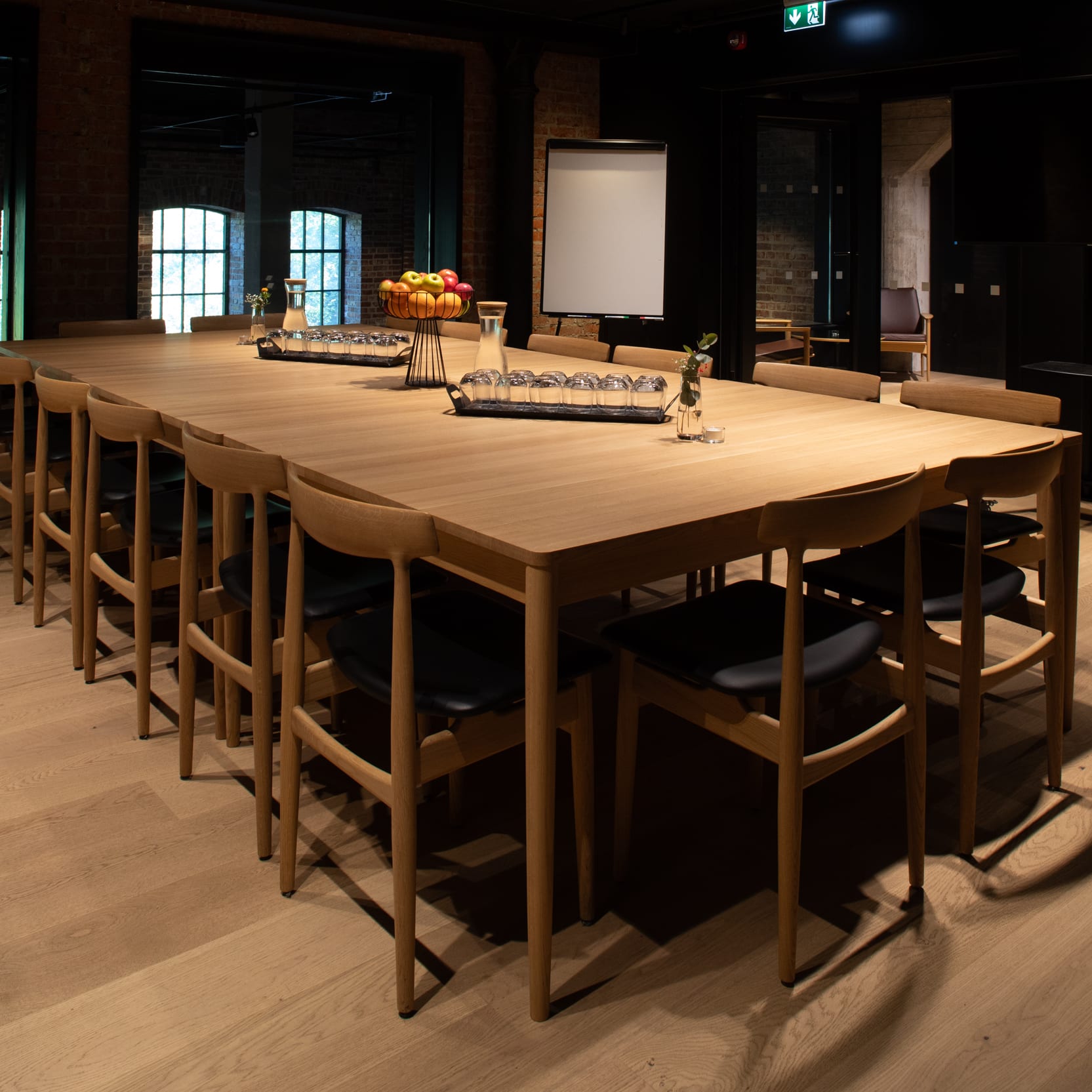A well-lit conference room features a long wooden table with chairs, refreshments, and a whiteboard. Windows provide natural light, with brick walls visible.