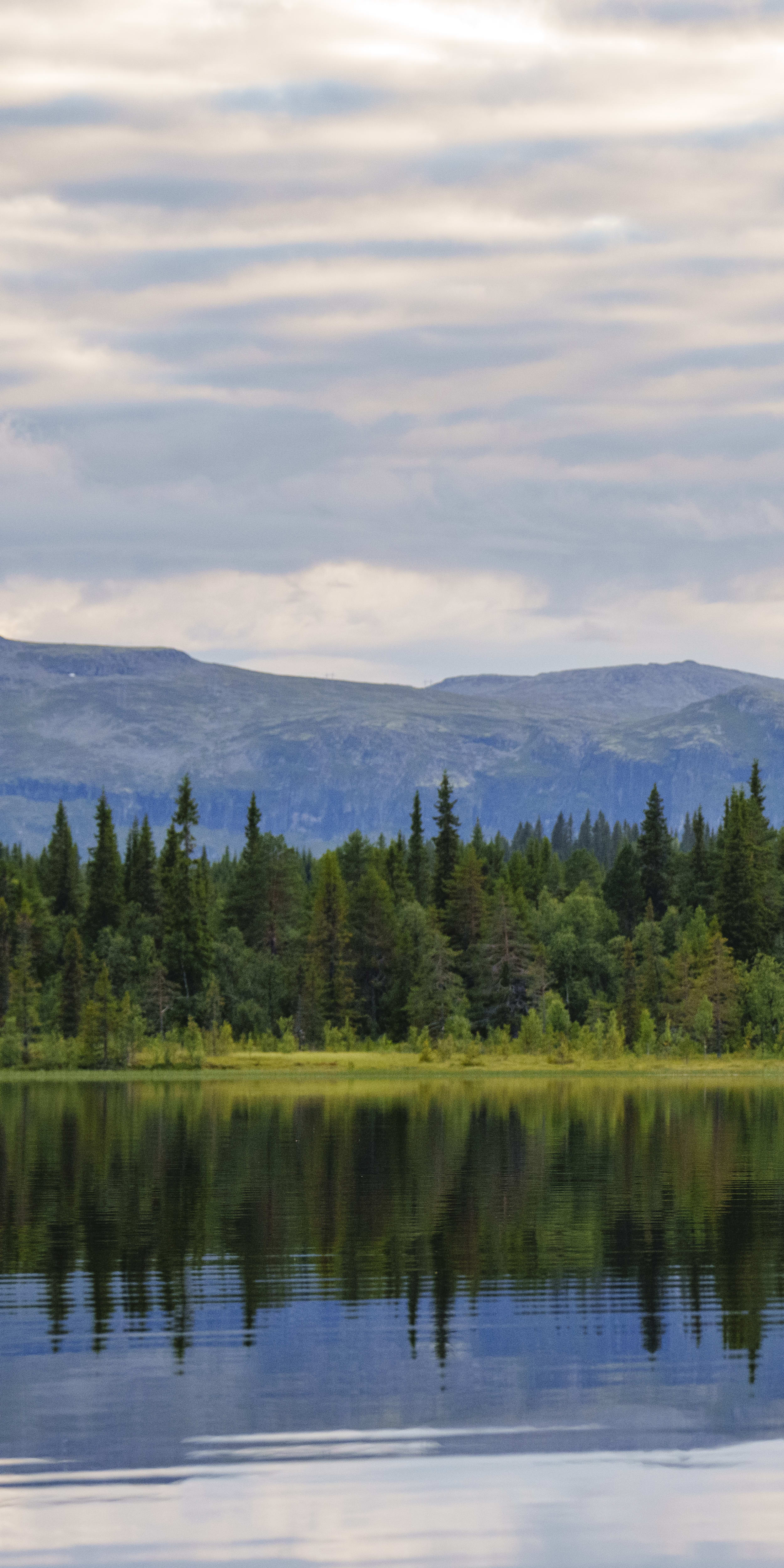 Two people in a canoe paddle across a calm lake. The lake reflects the sky, the clouds, and the forested mountain range in the background.  The people are wearing Norrøna clothing.
