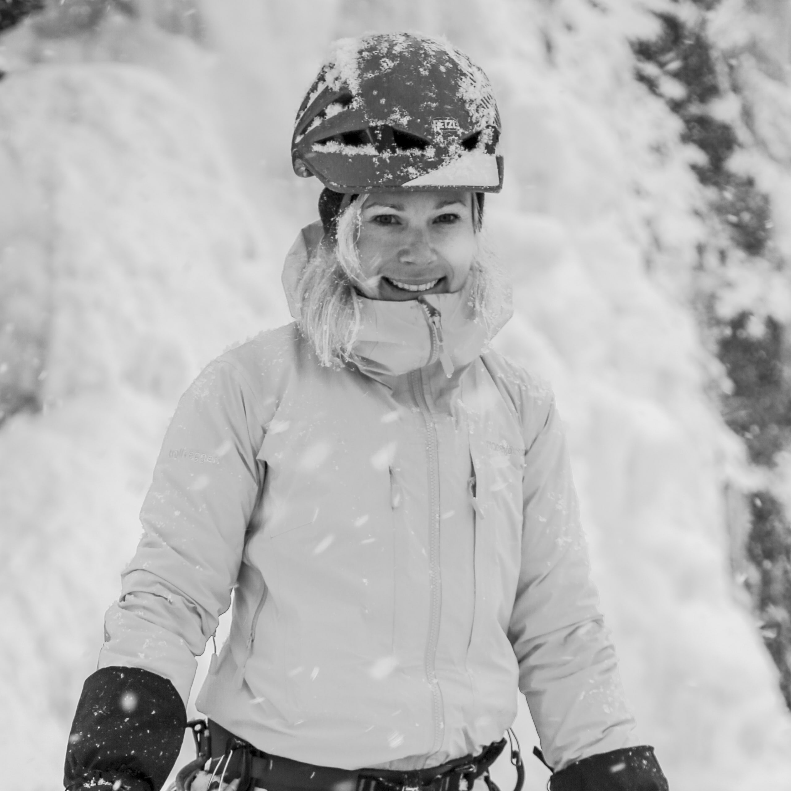 A woman wearing a Norrøna jacket and a helmet smiles for the camera. Snow falls around her.