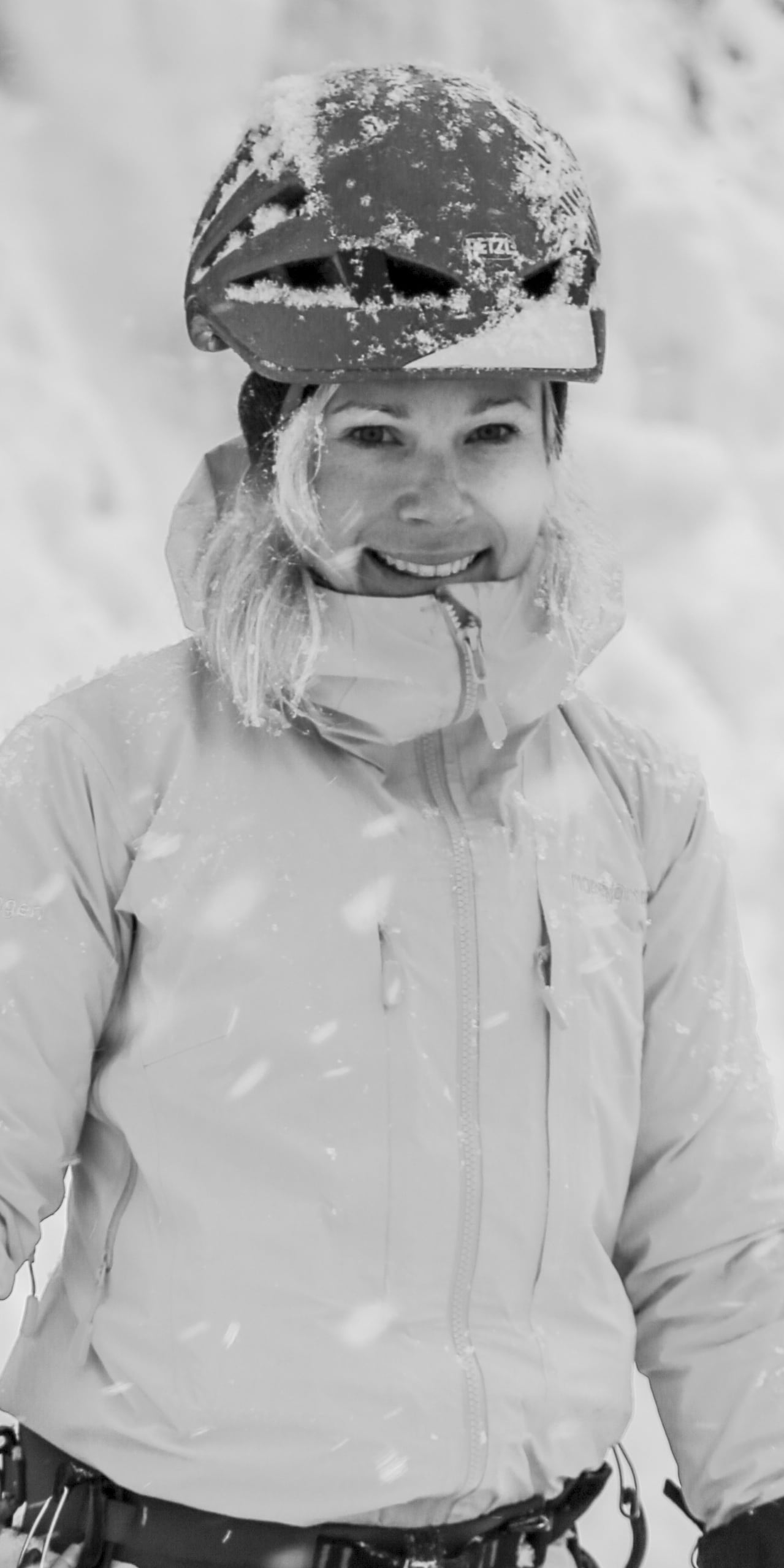 A woman wearing a Norrøna jacket and a helmet smiles for the camera. Snow falls around her. 
