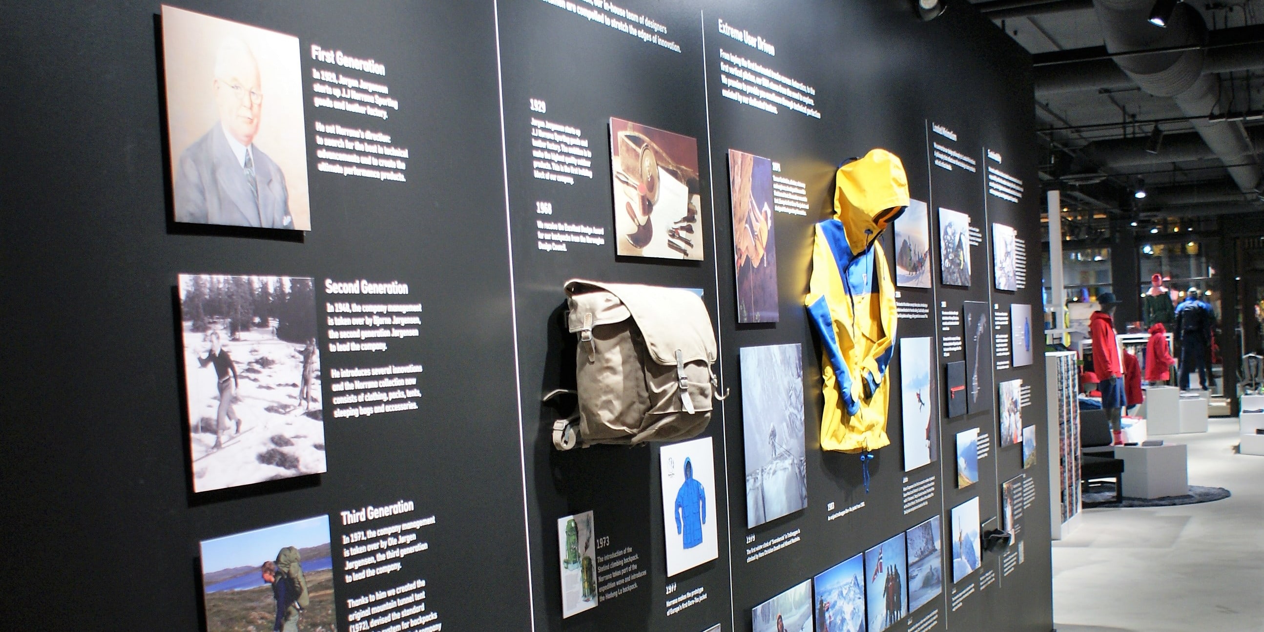 A black wall with photos showing the history of Norrøna, a Norwegian outdoor clothing company, from 1929 to present.  The photos show the company's founder, products, and people using the products.  There is a yellow Norrøna jacket on display.