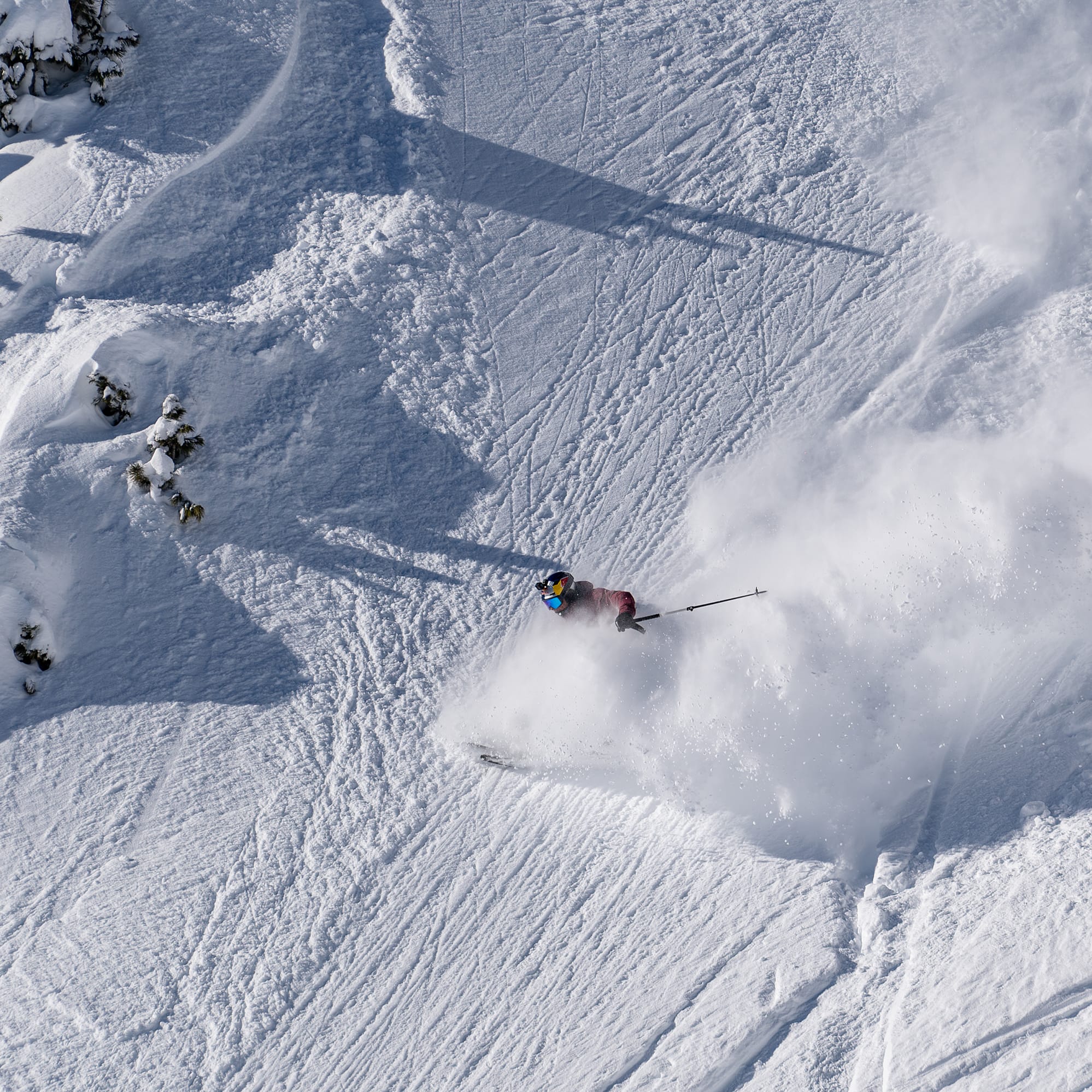 Aerial view of a skier in red Norrøna jacket, amidst snow spray, skiing down a steep, snowy mountain slope.