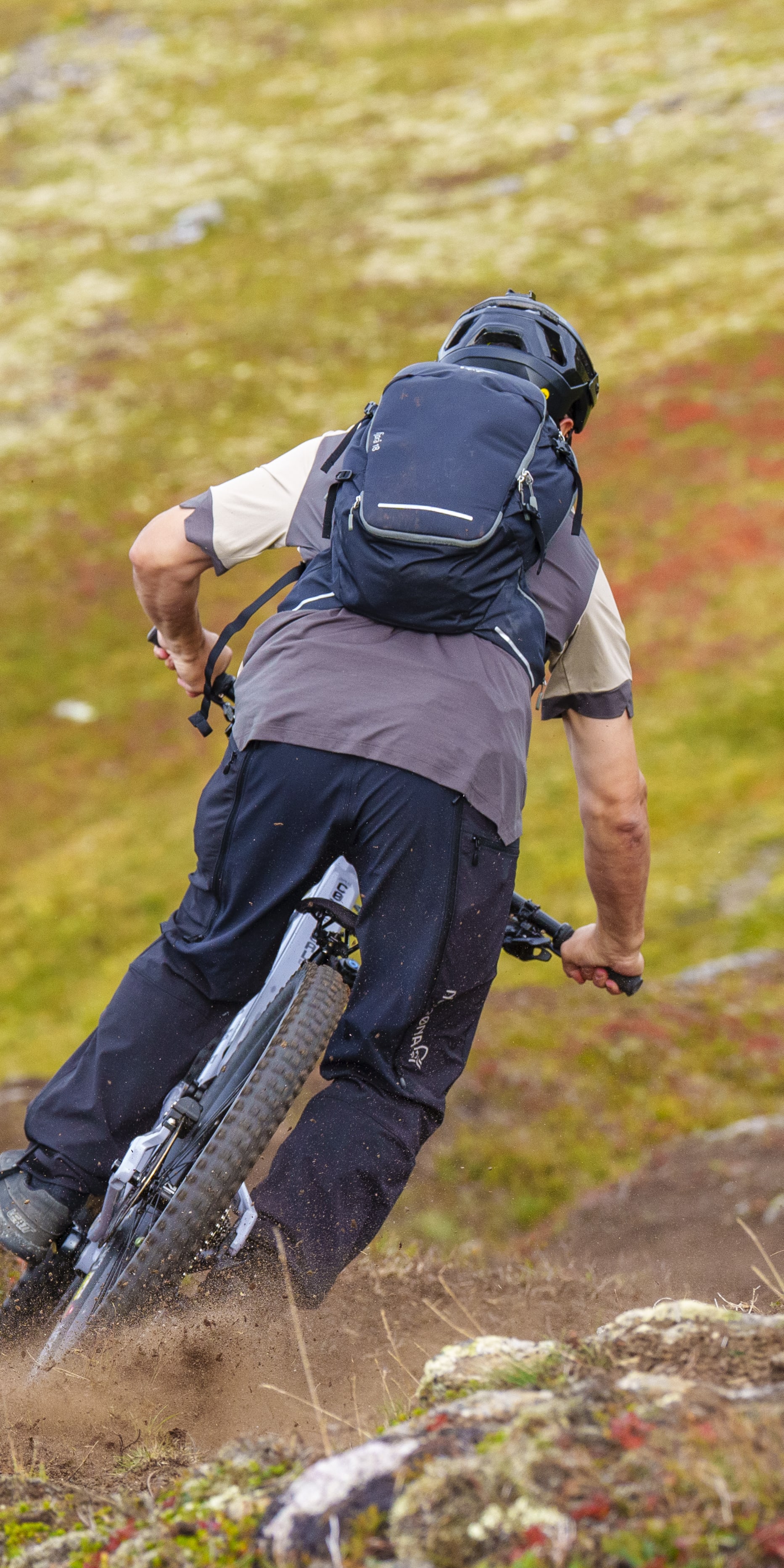 A mountain biker navigates a dirt trail, kicking up dust. They wear a helmet, backpack, and Norrøna pants. Back view.

