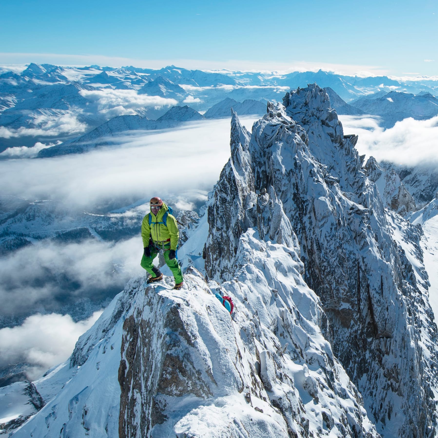 Two mountaineers ascend a snow-covered peak on a clear day, wearing Norrøna outerwear.  Mountains and clouds fill the background.
