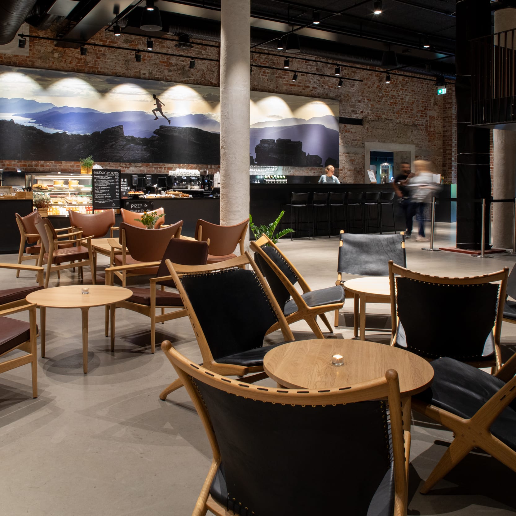 Interior of a cafe with tables and chairs. A scenic mural is on the wall. A bar and stairwell are in the background.