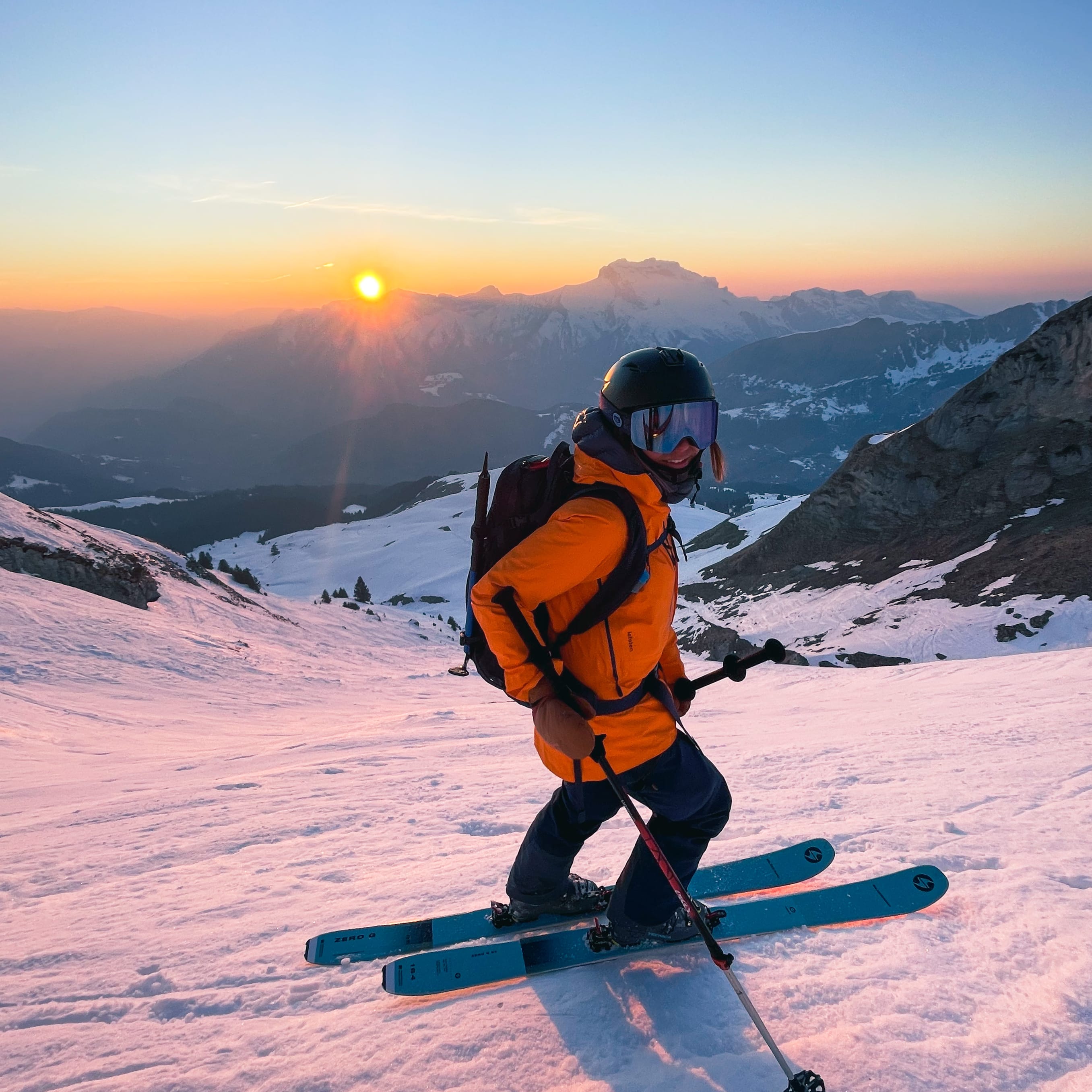 A skier in Norrøna outerwear ascends a snowy mountain at sunrise. Mountains are in the background.