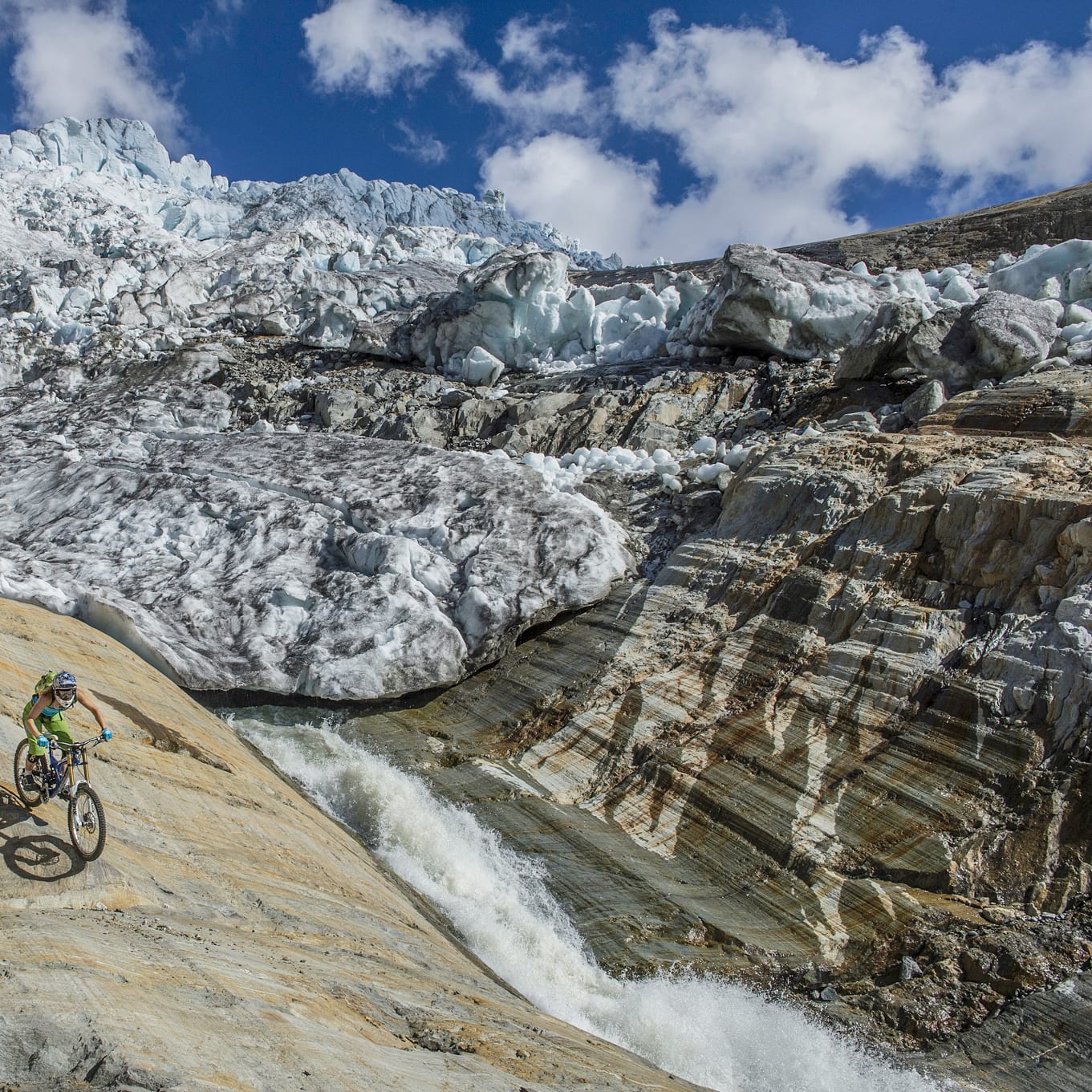 A mountain biker in Norrøna clothing rides across a striated rock face with a waterfall and glacier backdrop under a blue sky.