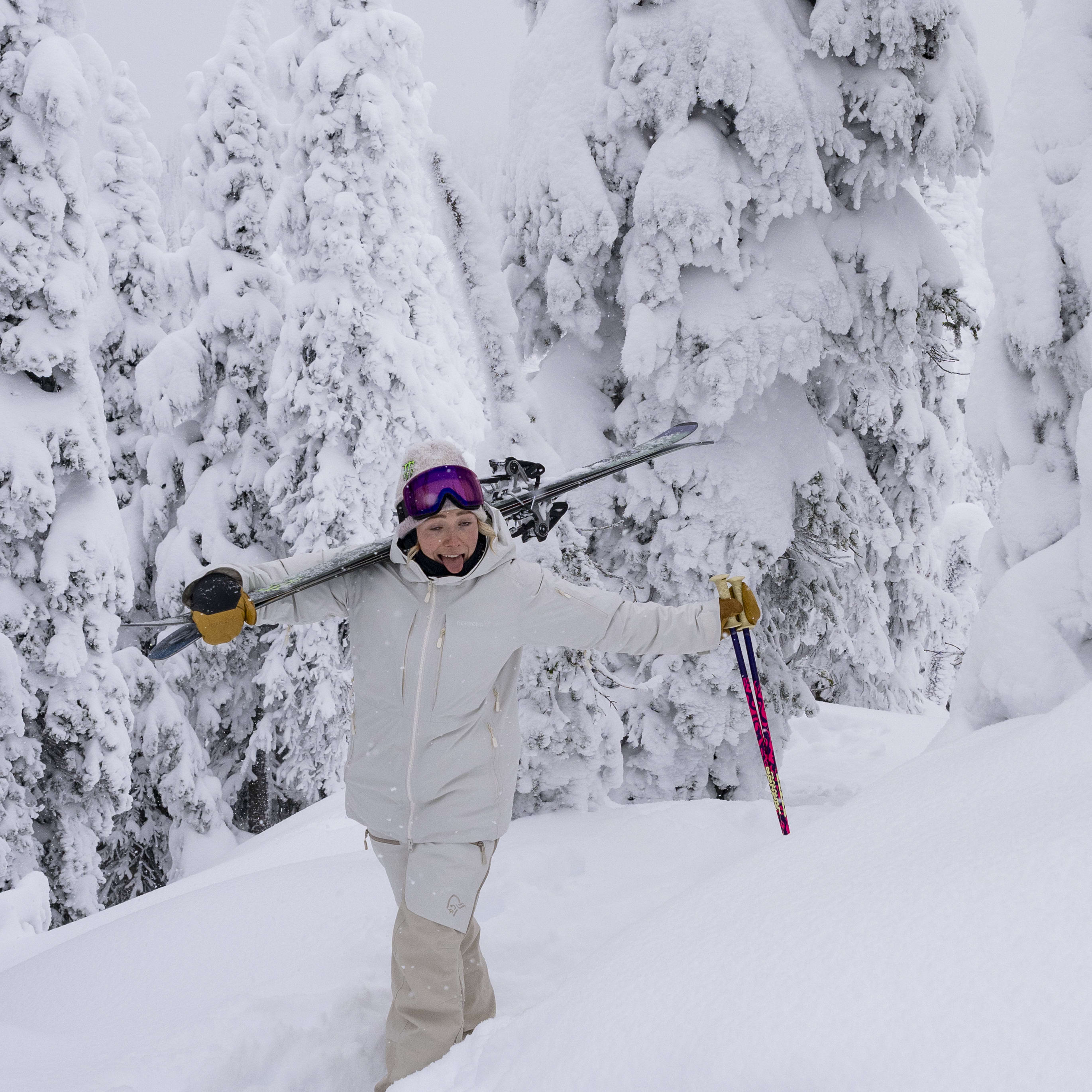 A woman in a Norrøna jacket and pants, skis over her shoulder, smiles in a snowy forest.