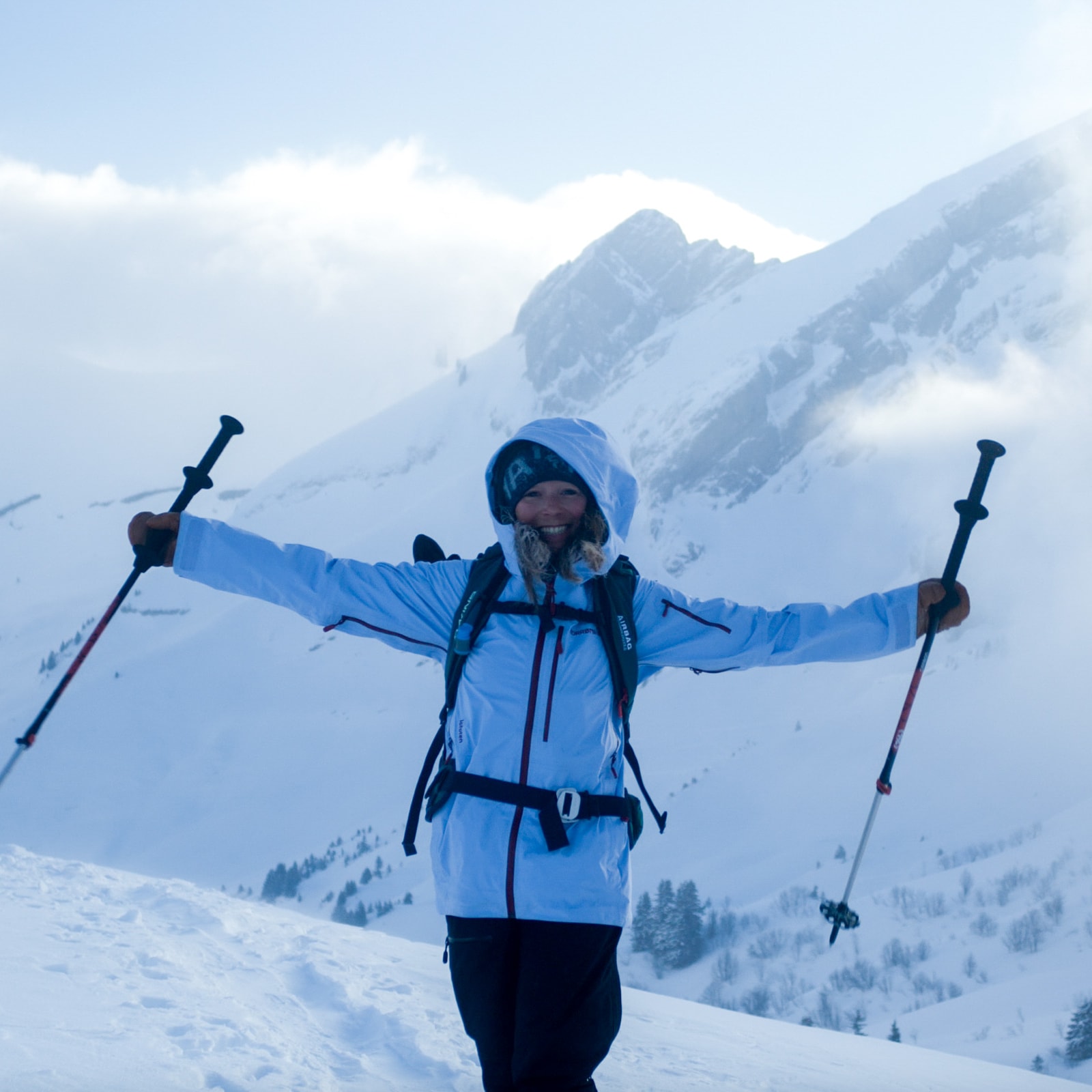 Woman hiking on snow mountain, arms outstretched, holding ski poles. She wears a Norrøna jacket and backpack. Fog and mountain in background.