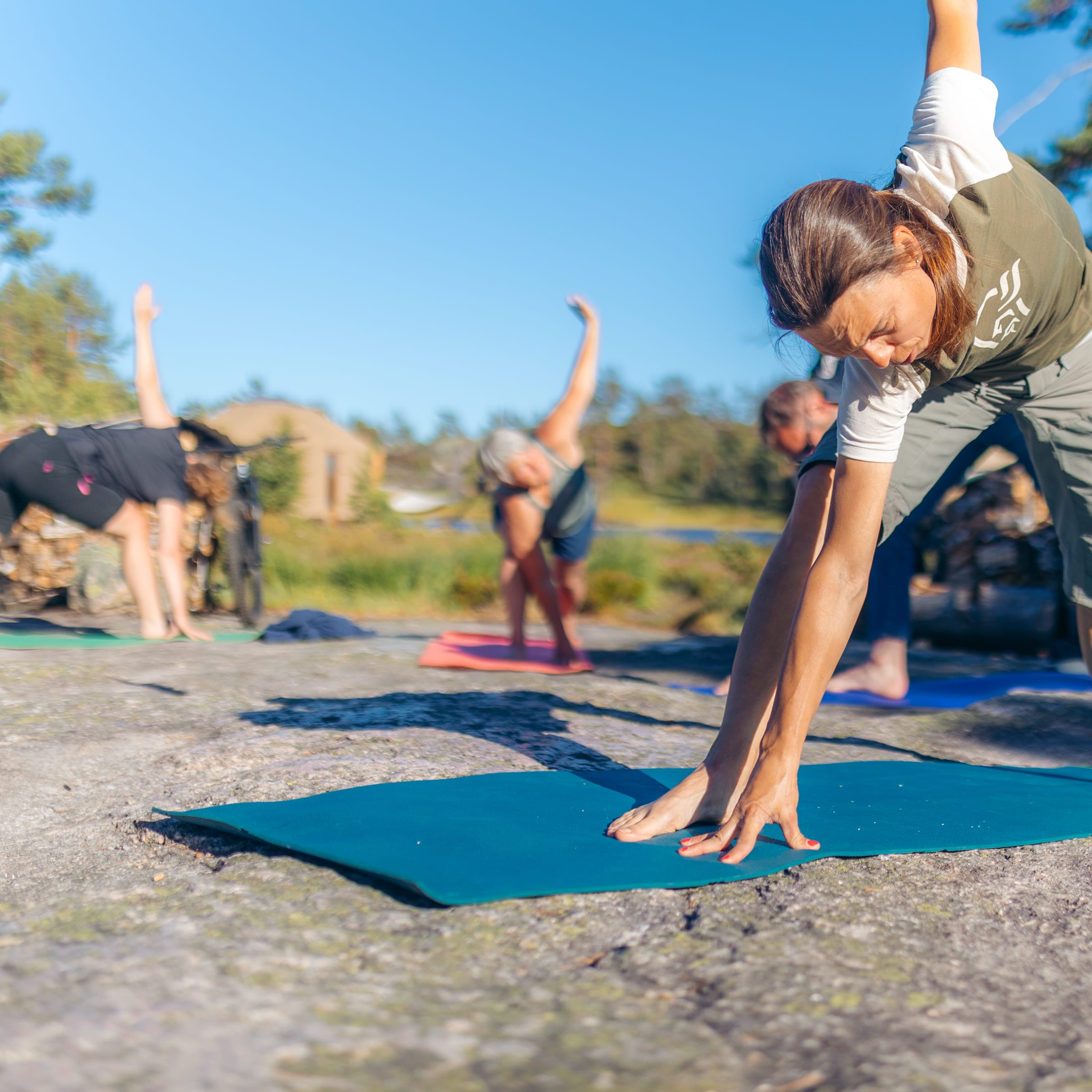A woman wearing a Norrøna shirt and pants performs yoga on a blue mat outdoors. Two other people are visible in the background. 
