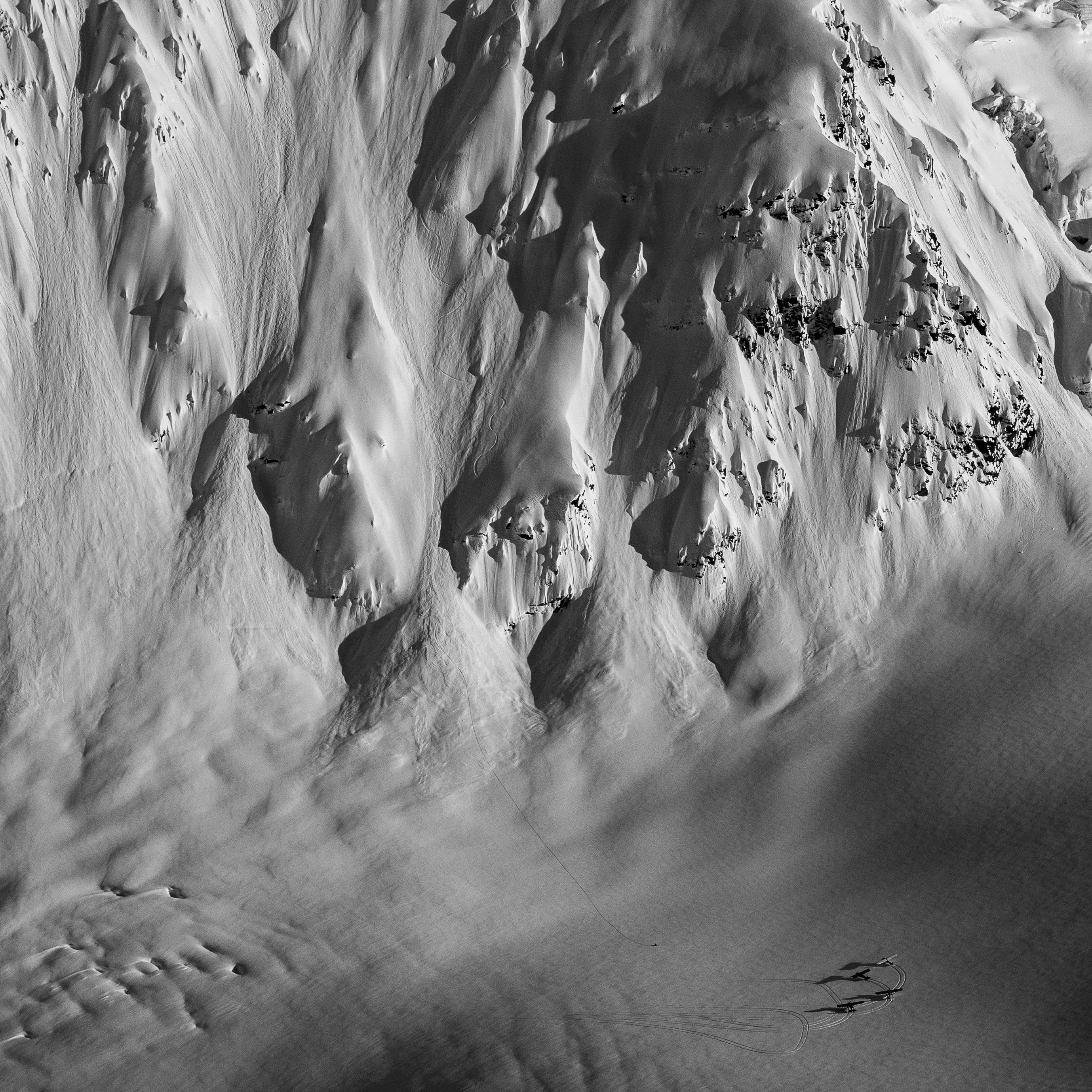 Monochrome aerial shot of a snow-covered mountain face with ski tracks. Small figures are visible at the bottom.
