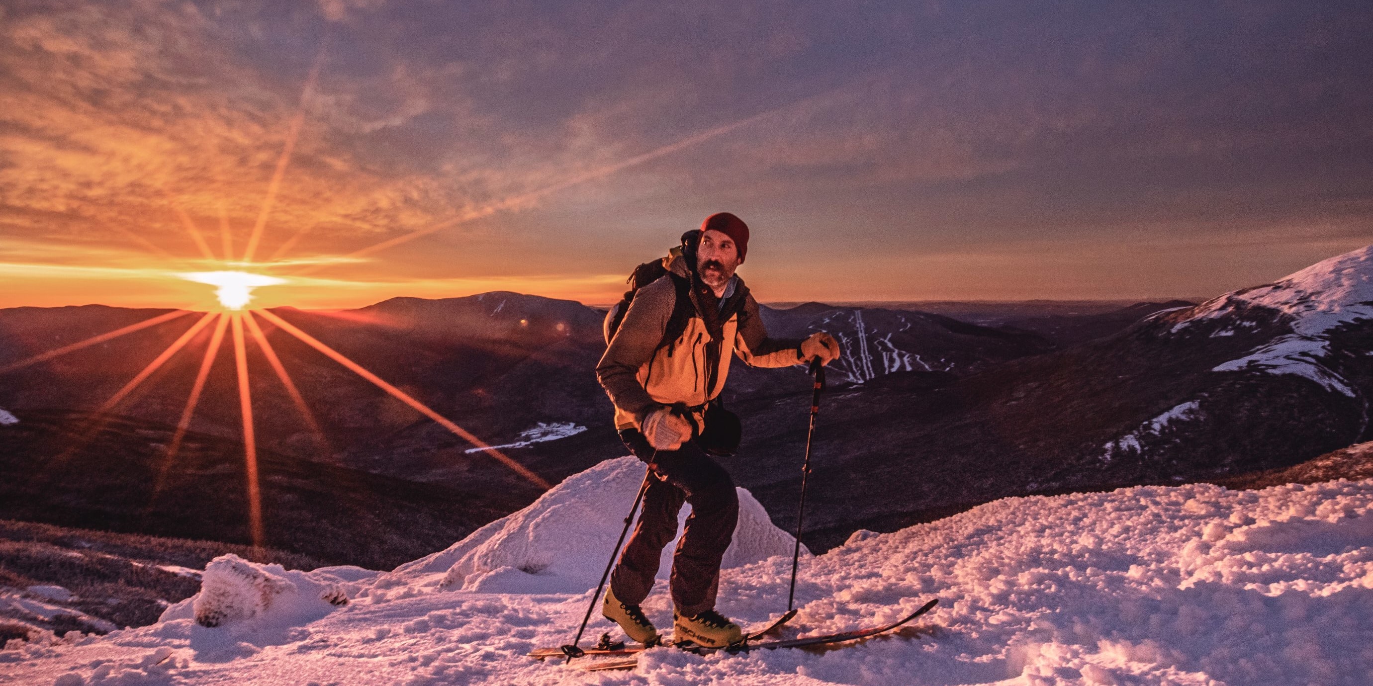 A man wearing a Norrøna jacket skis down a snowy mountain with a stunning sunrise in the background. 
