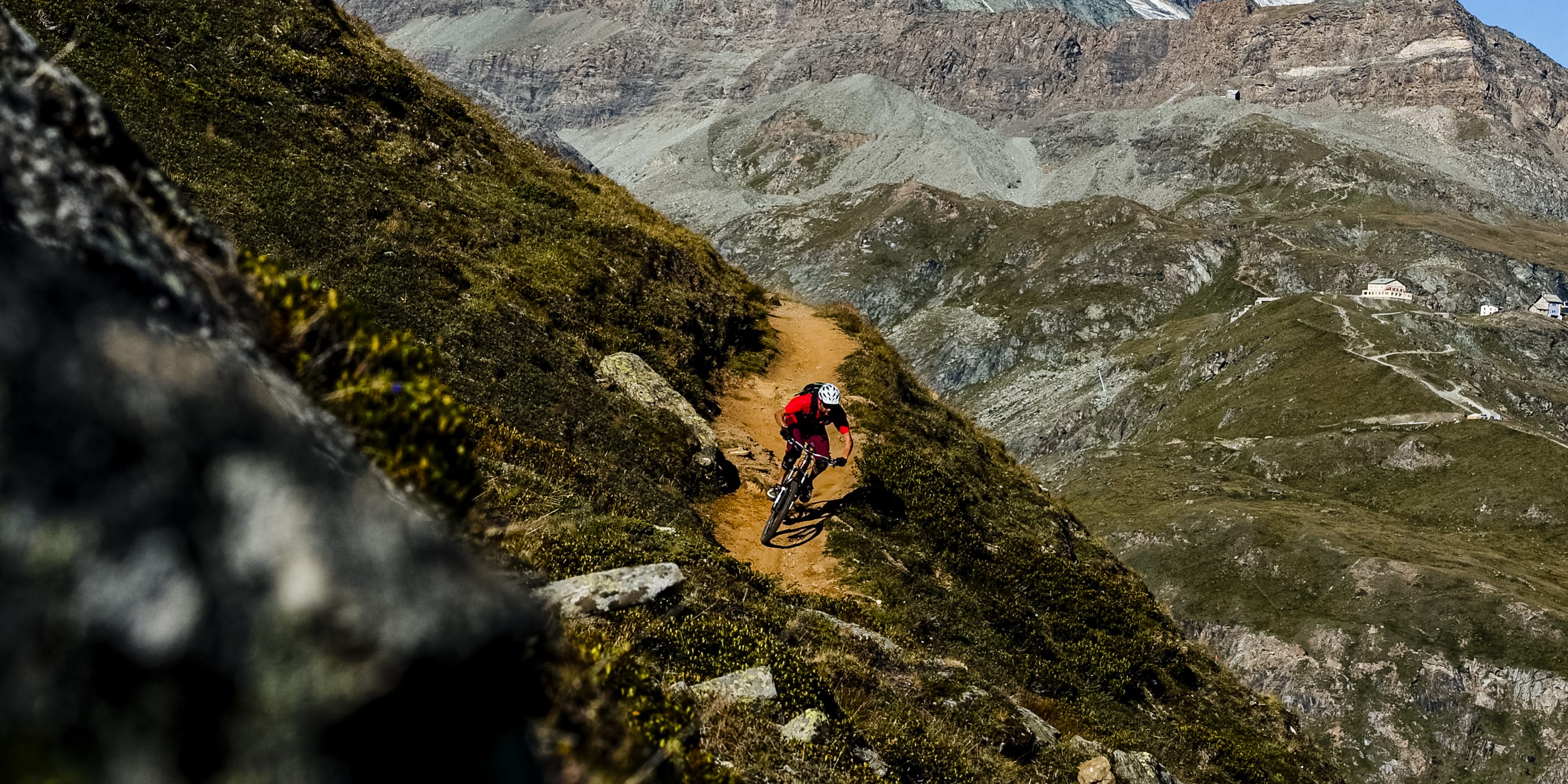 A person wearing Norrøna gear mountain bikes down a dirt trail with the Matterhorn in the background. 
