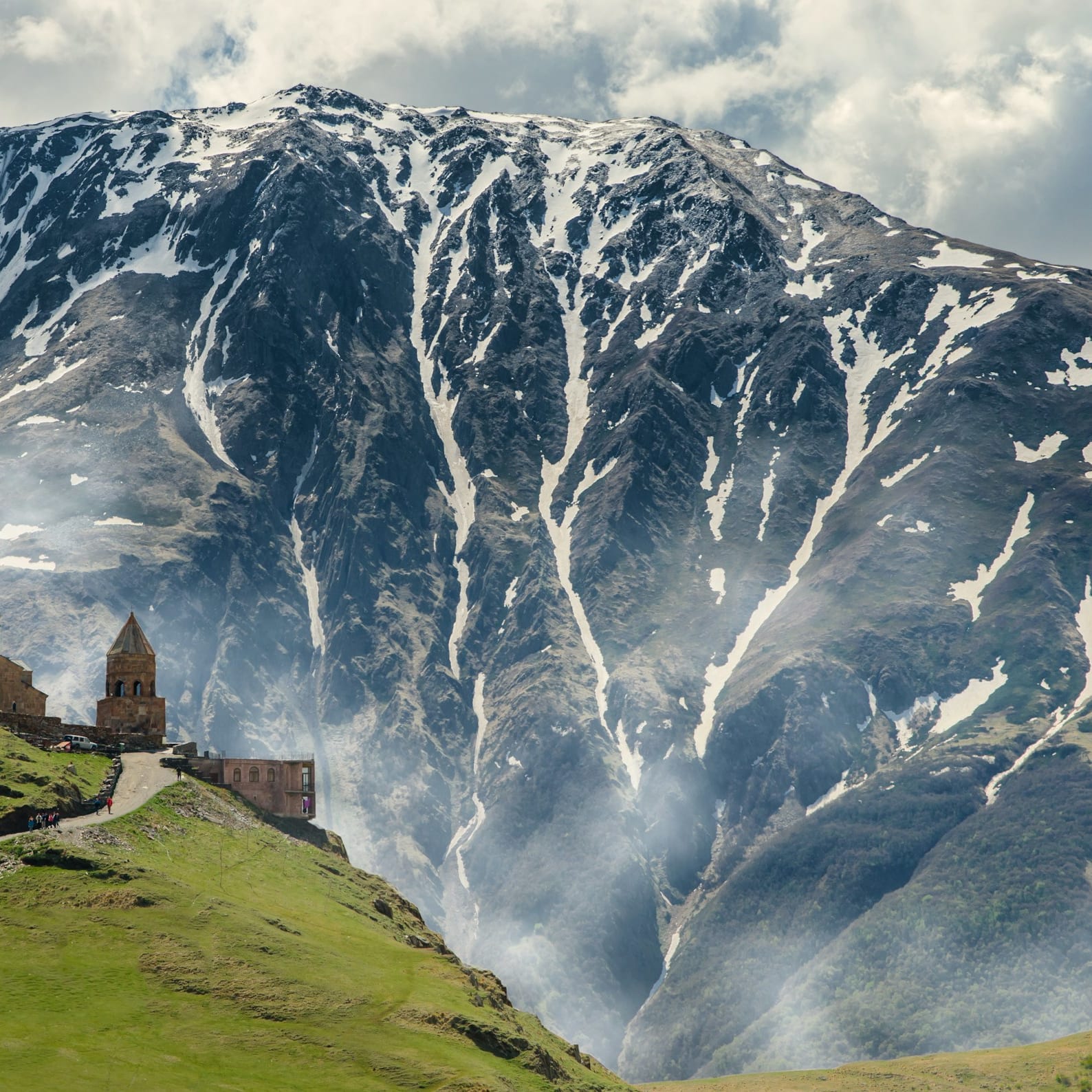 Gergeti Trinity Church on a green hill with a massive, snow-streaked mountain in the background.