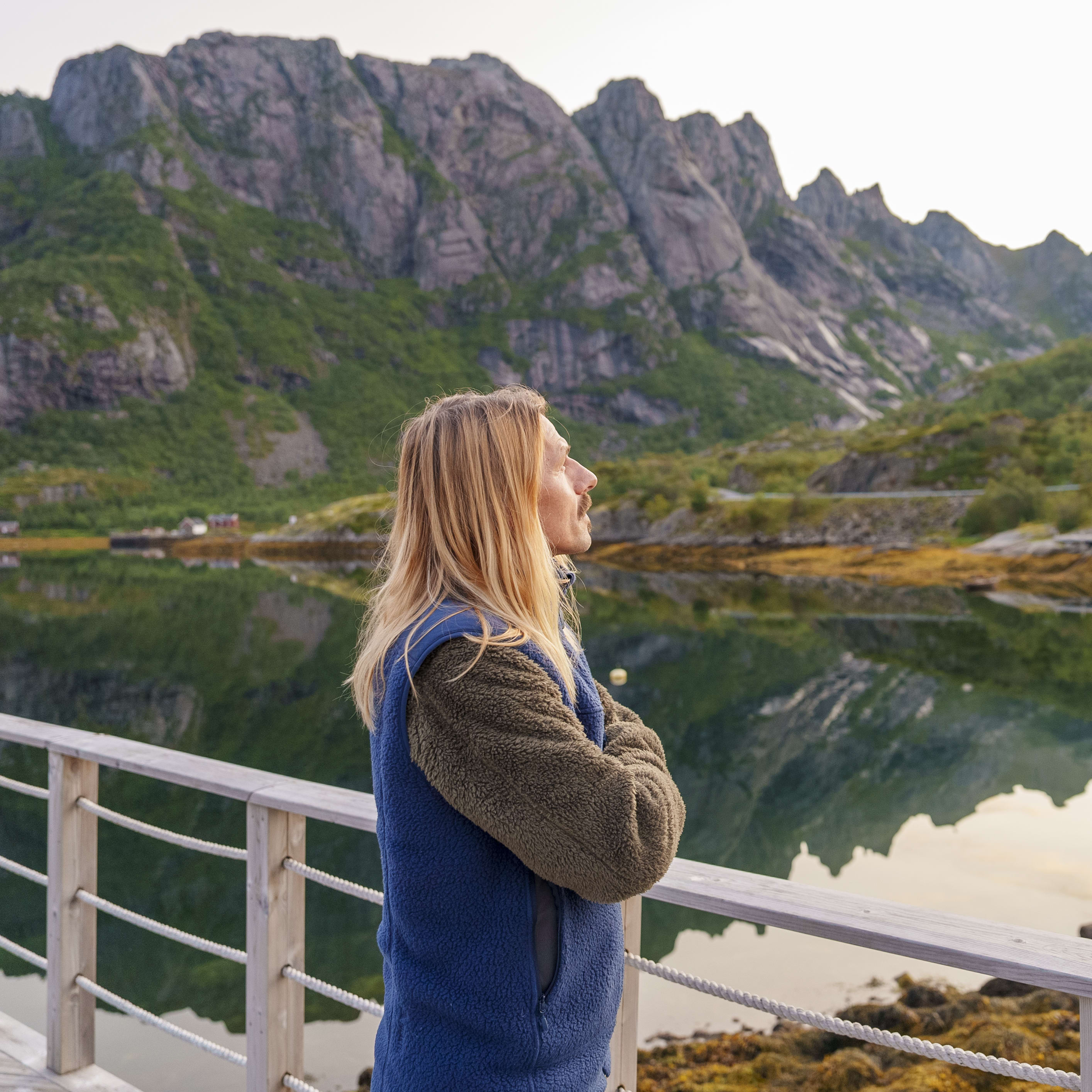 A person with long blonde hair wearing a Norrøna jacket stands on a wooden railing overlooking a calm body of water. Mountains and green hills are reflected in the water in the background.