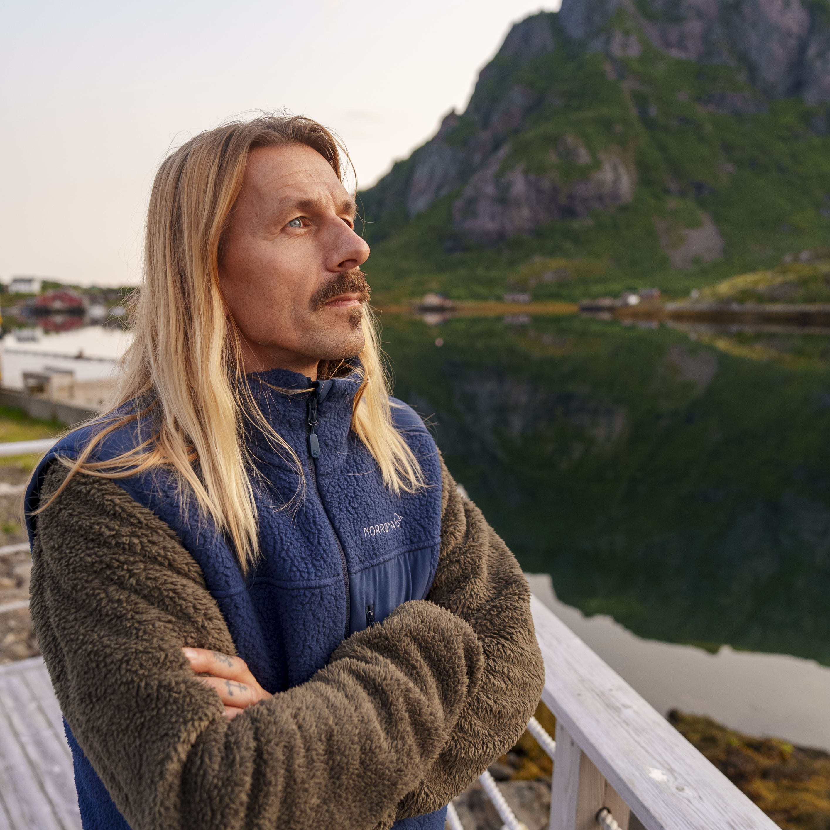 Man with long blonde hair wearing a blue Norrøna fleece jacket, standing on a wooden pier, looking out over a body of water and mountains. 
