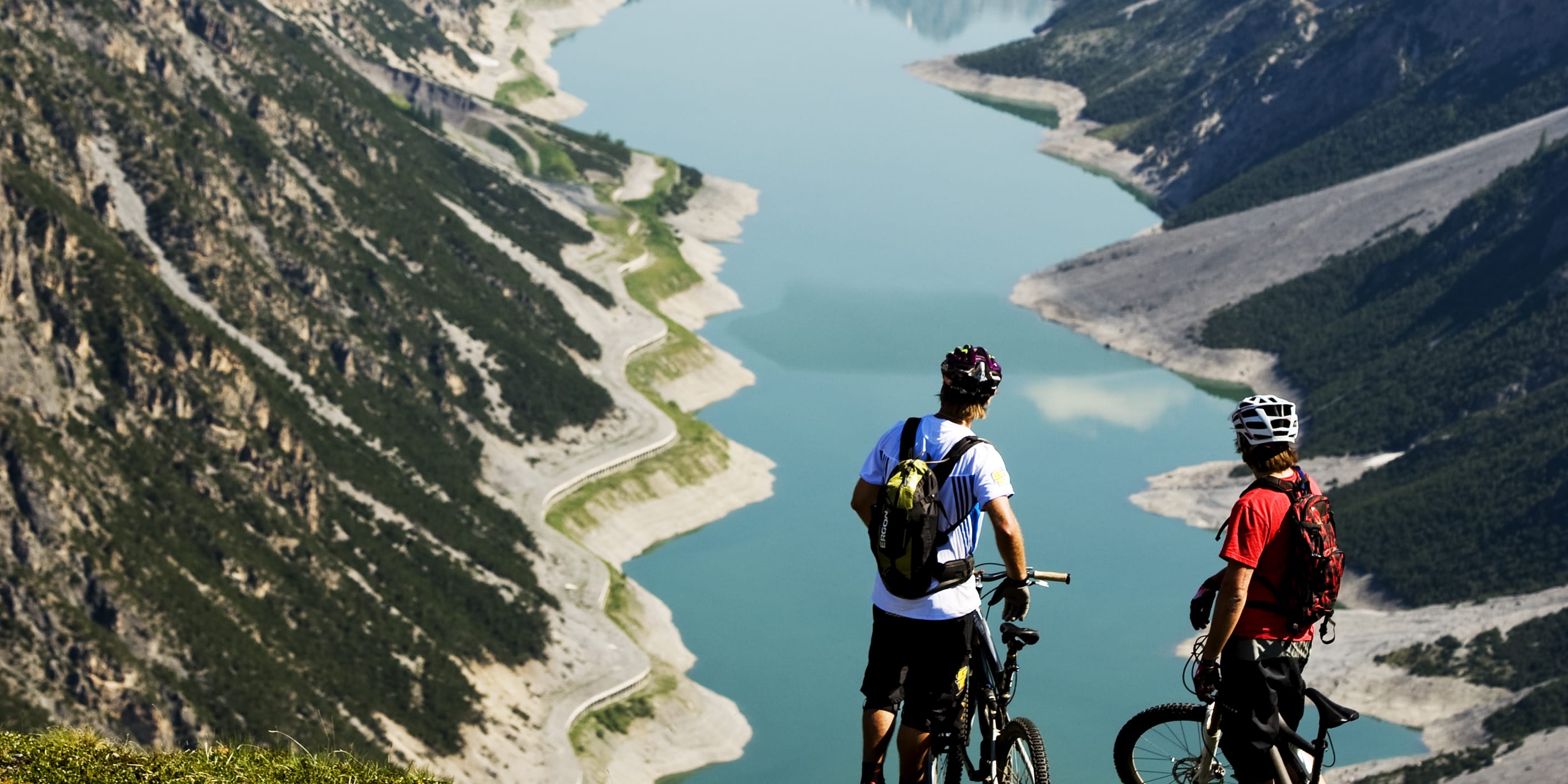 Two mountain bikers stand on a grassy hill overlooking a lake and mountains. They are wearing helmets and Norrøna backpacks.  
