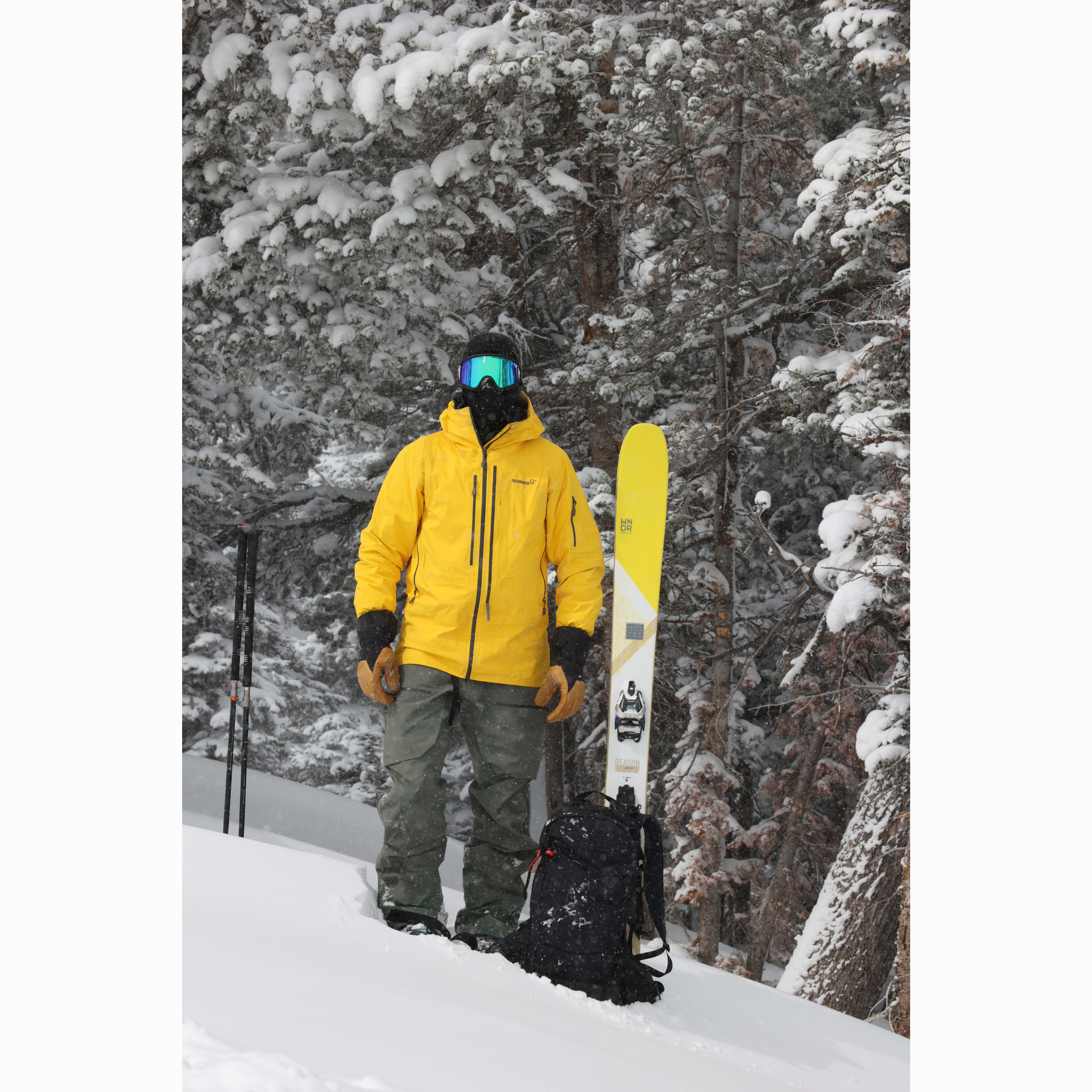 A skier in a yellow Norrøna jacket, gray pants, and goggles stands in a snowy forest next to skis and a backpack.