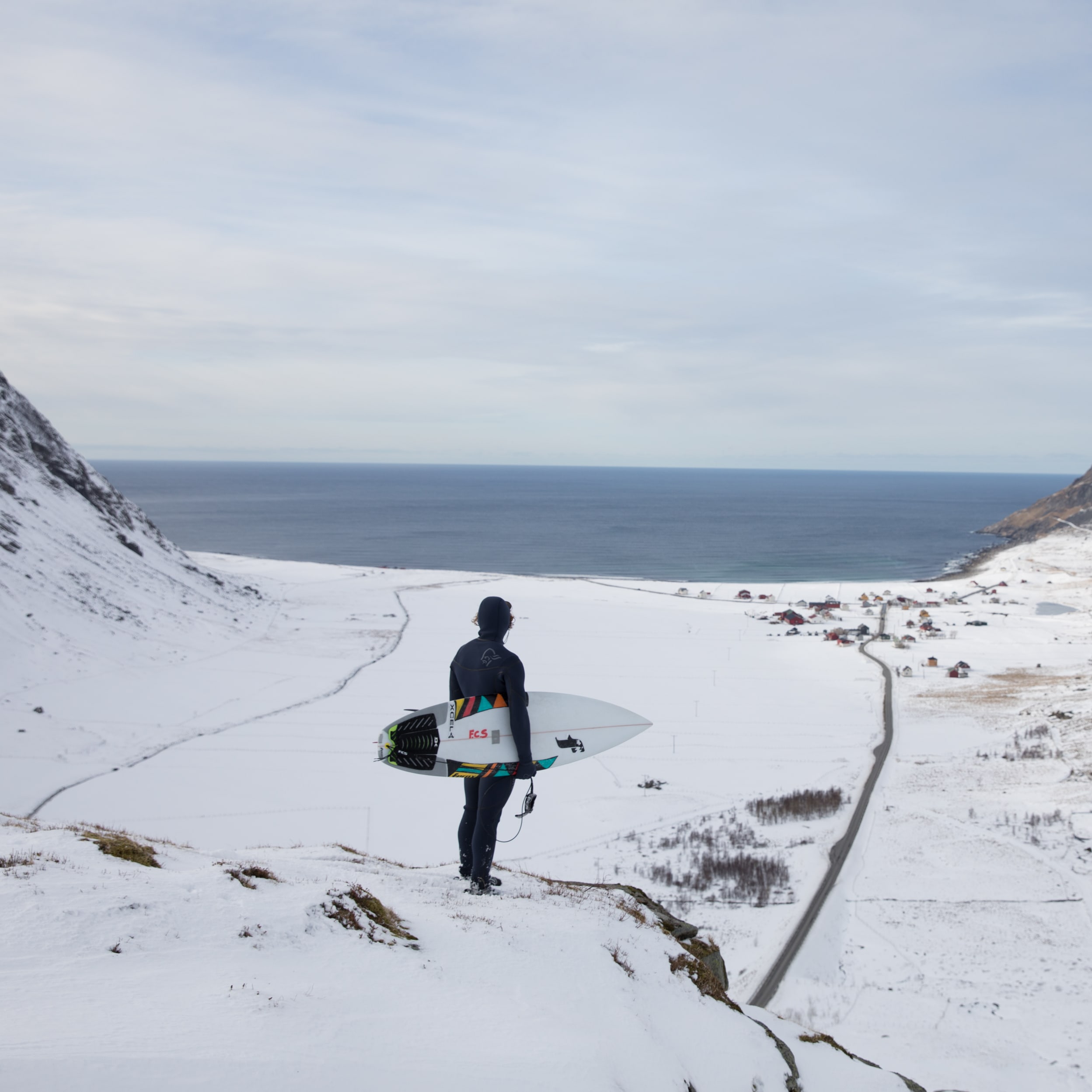 A person wearing a Norrøna suit and holding a surfboard, standing on a snowy mountain overlooking a town and a body of water. 
