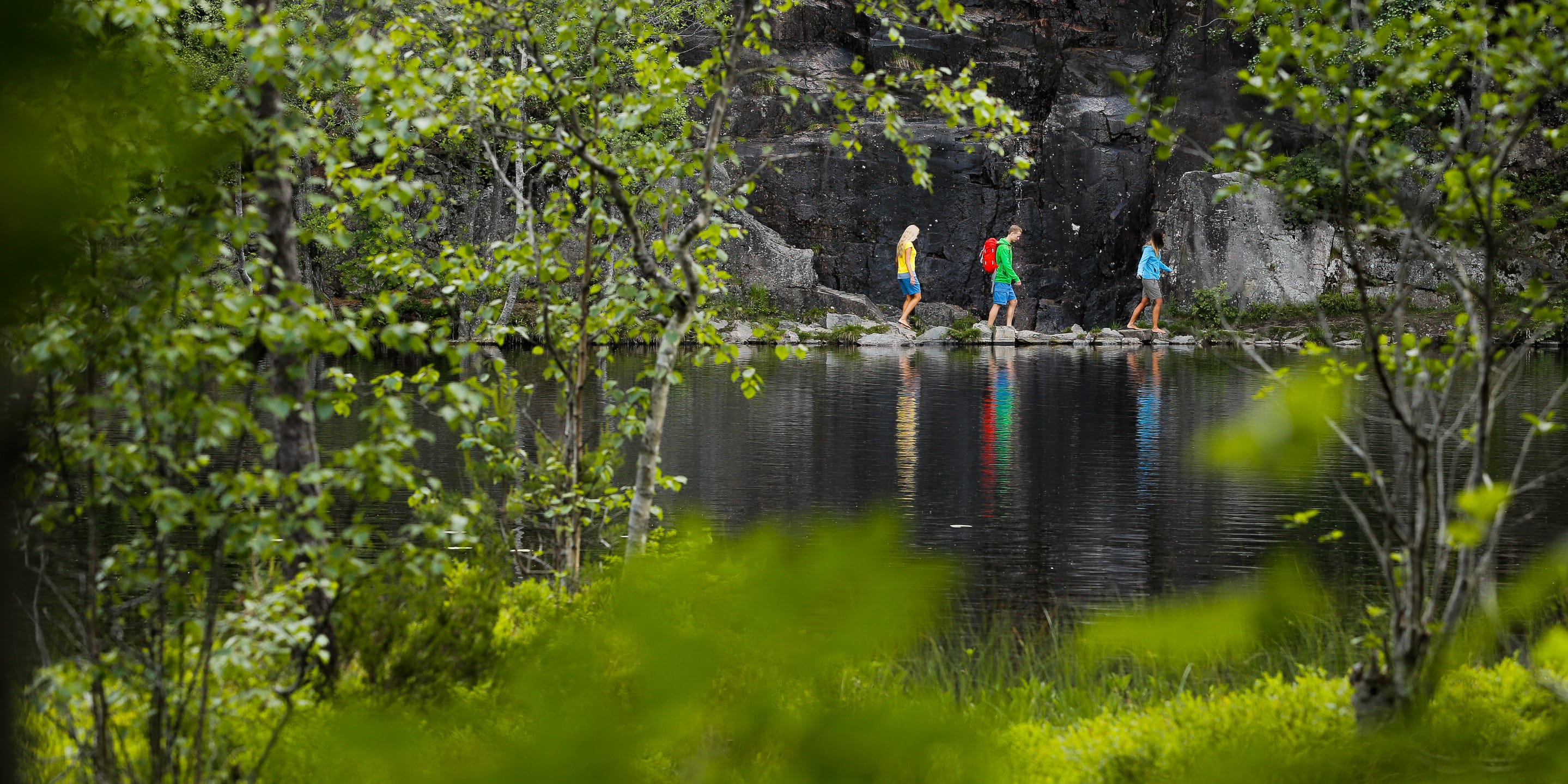 Three people wearing Norrøna clothing walk on rocks near a lake. The lake and a rocky cliff are in the background. Green foliage obscures the view.
