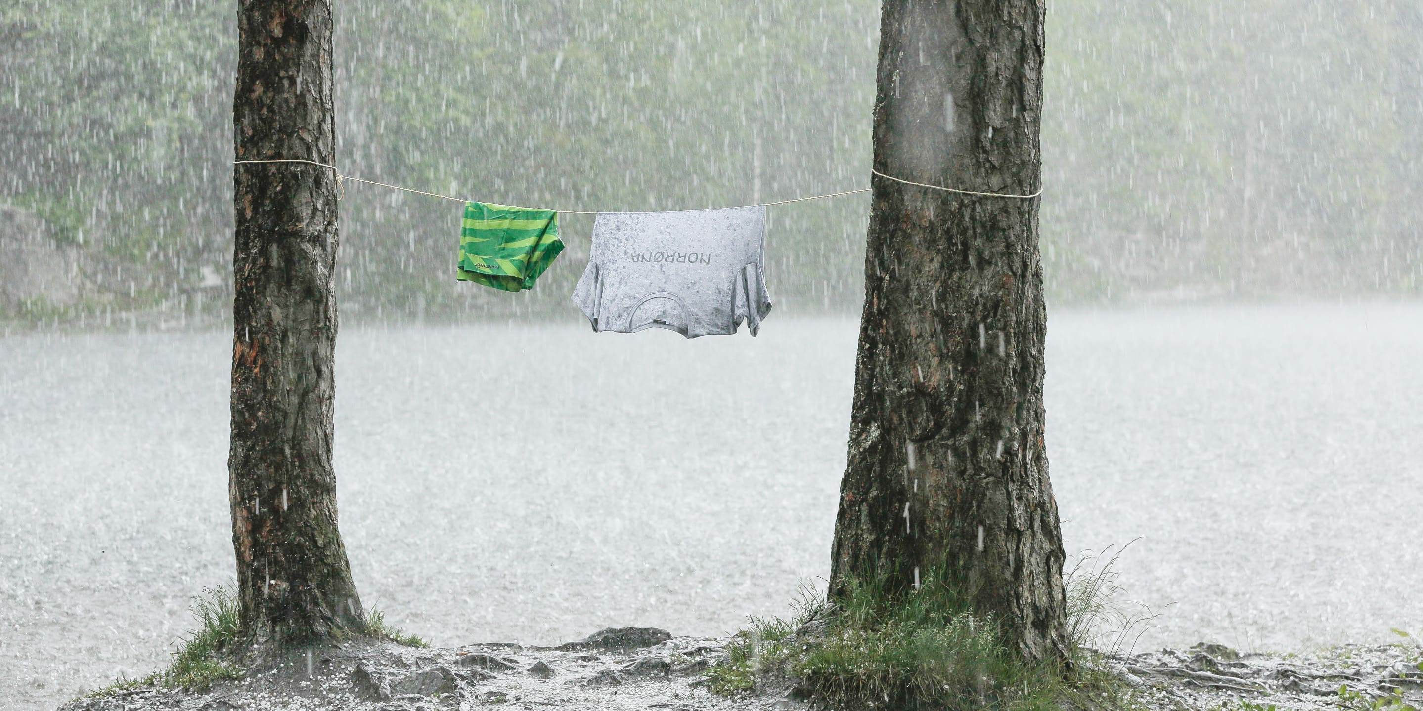 Two trees with a Norrøna shirt and green and white striped shirt hanging on a line in front of a lake. It is raining heavily.